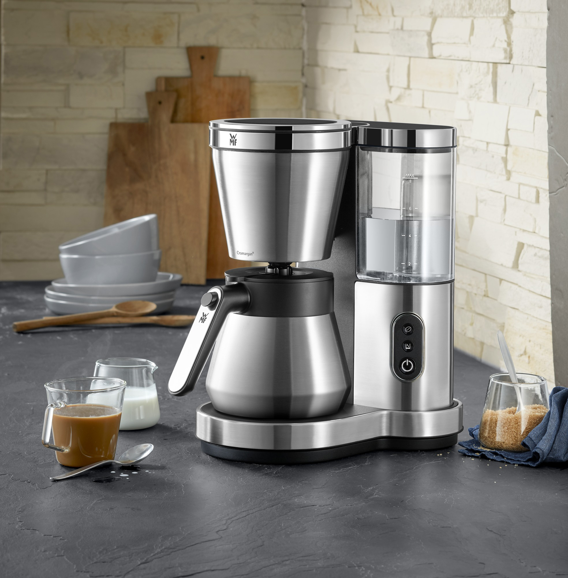 Koffiezetapparaat met chromen en zwarte afwerking, een kopje koffie en ingrediënten op een grijs aanrecht.