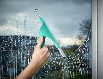 Eine Hand verwendet einen grünen Fensterabzieher, um ein Fenster zu reinigen. Wasser und ein verschwommener Hintergrund.