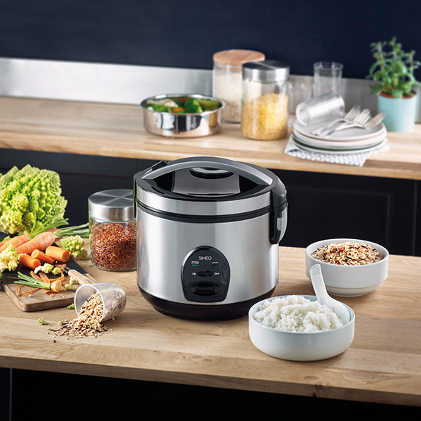 Rice cooker with food items, vegetables, and utensils on a wooden surface.