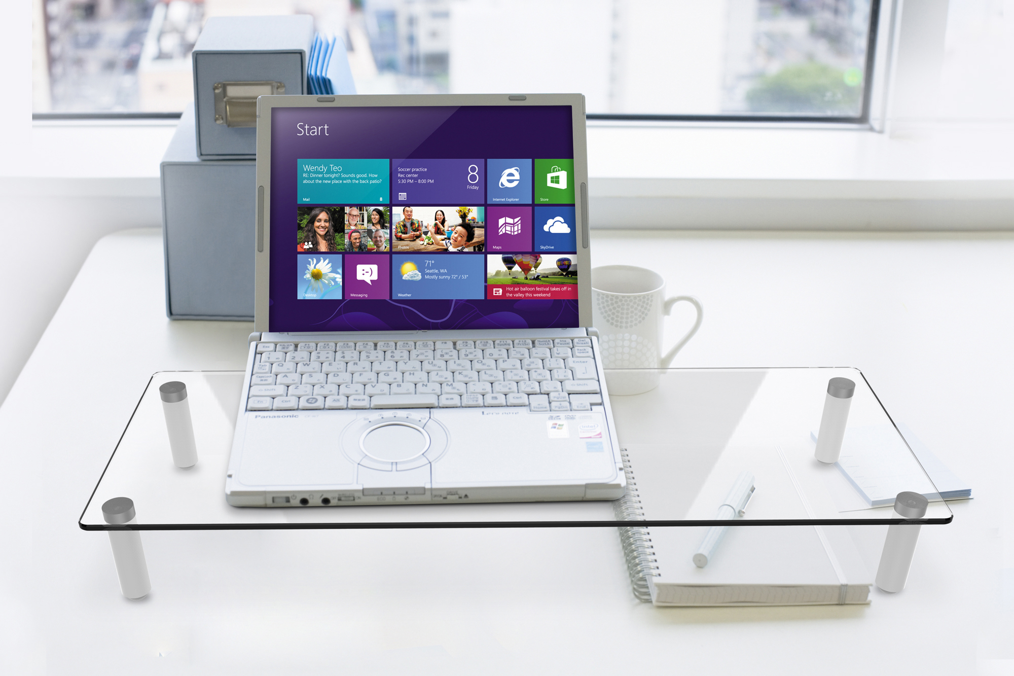 Ein Laptop auf einem Glastisch. Ein Monitor, eine Tasse und Bürobedarf sind ebenfalls sichtbar.