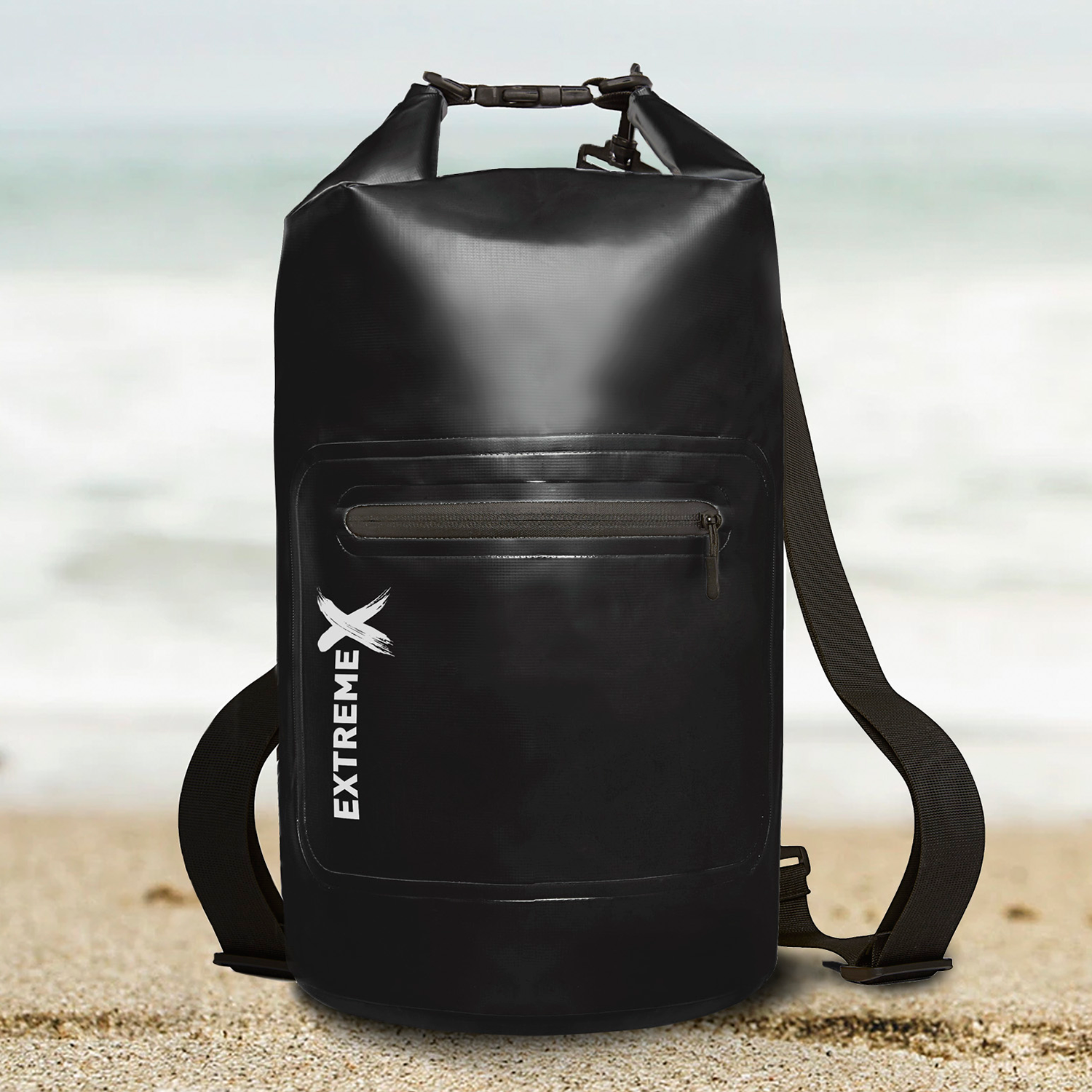 A black waterproof backpack stands on sand, with a beach and ocean background. It has a white logo and a shoulder strap.