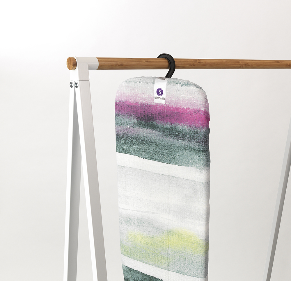 Een kleurrijke strijkplankhoes hangt aan een kledingrek. Het heeft een zwarte hanger. De hoes heeft roze, groene en witte strepen.