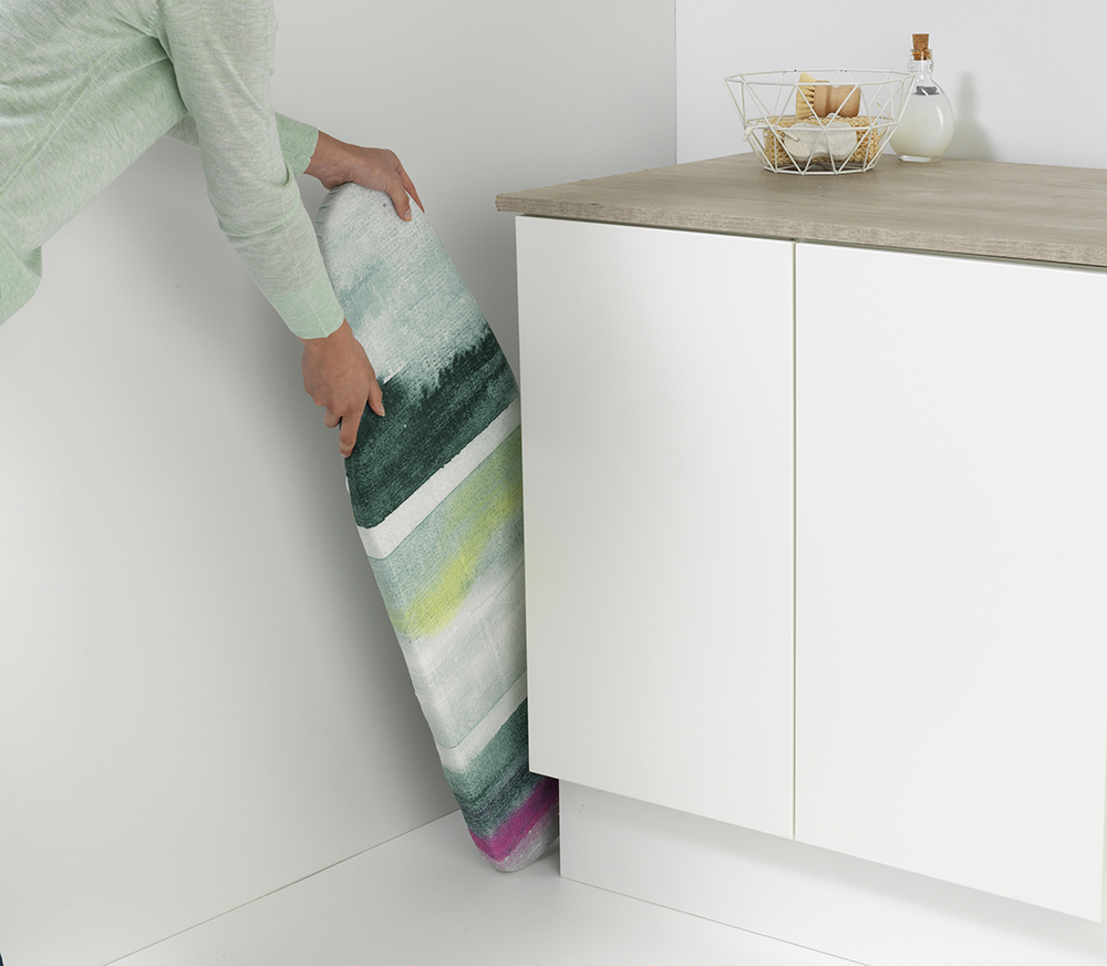 Een persoon plaatst een kleurrijk vloerkleed in een kast. Het vloerkleed heeft groene, witte en roze strepen. Een wastafel en kast zijn op de achtergrond.
