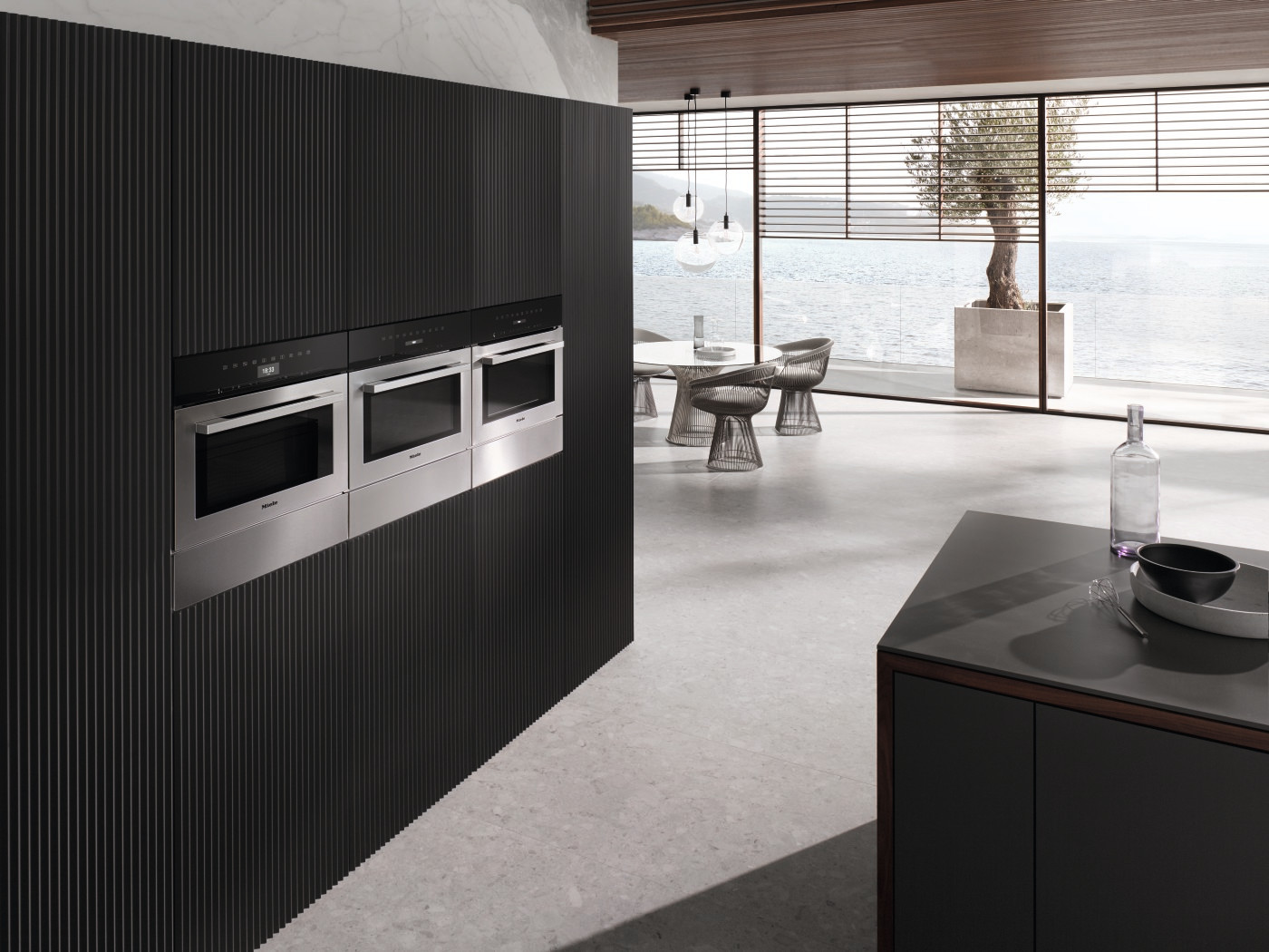 Moderne keuken met geïntegreerde ovens in een donkere houten wand. Open naar een eethoek en uitzicht op zee.