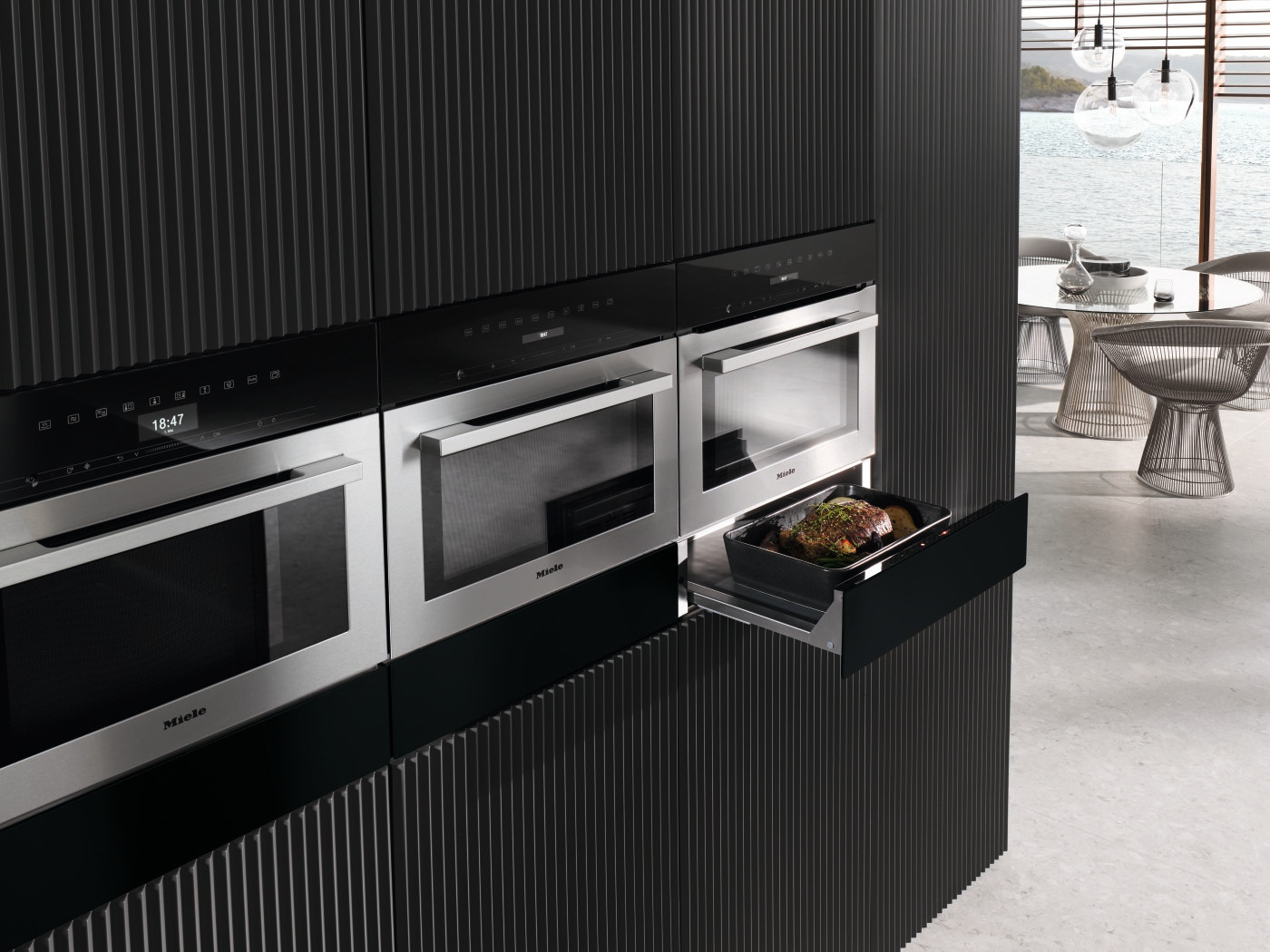 Keuken met zwarte kasten, roestvrijstalen apparaten, open ovenschuif met eten, lichte achtergrond.