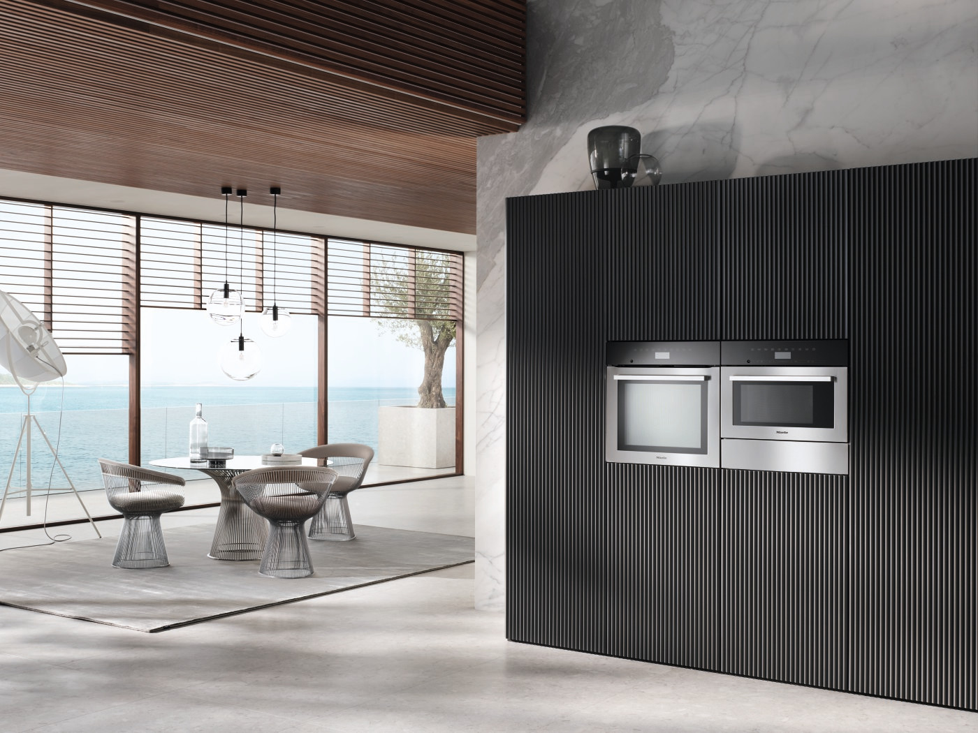 Modern interior with a dining set, a black wall with built-in ovens and a view of the sea.