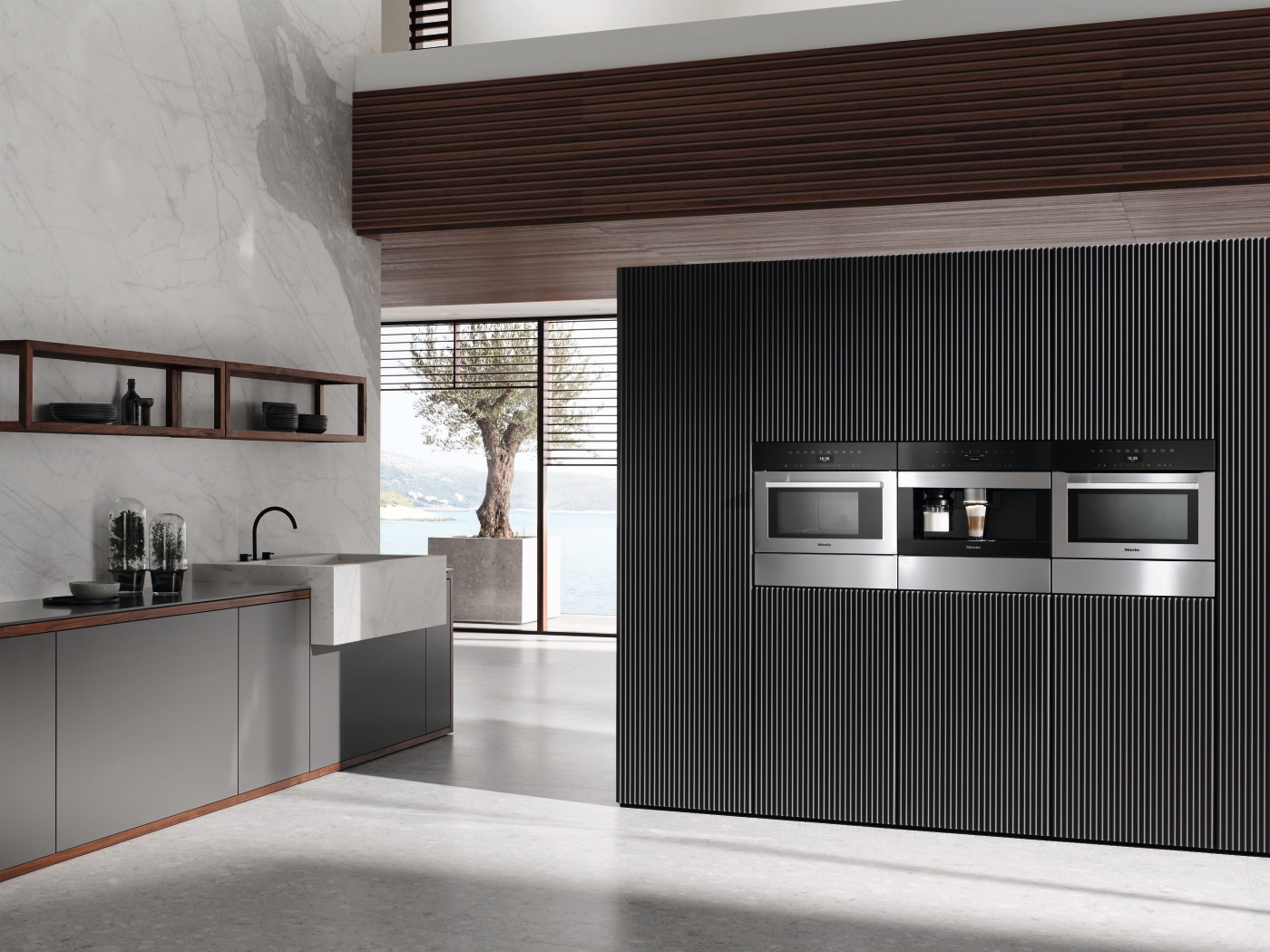 A modern kitchen interior with built-in appliances, a grey island and a large window overlooking a body of water.