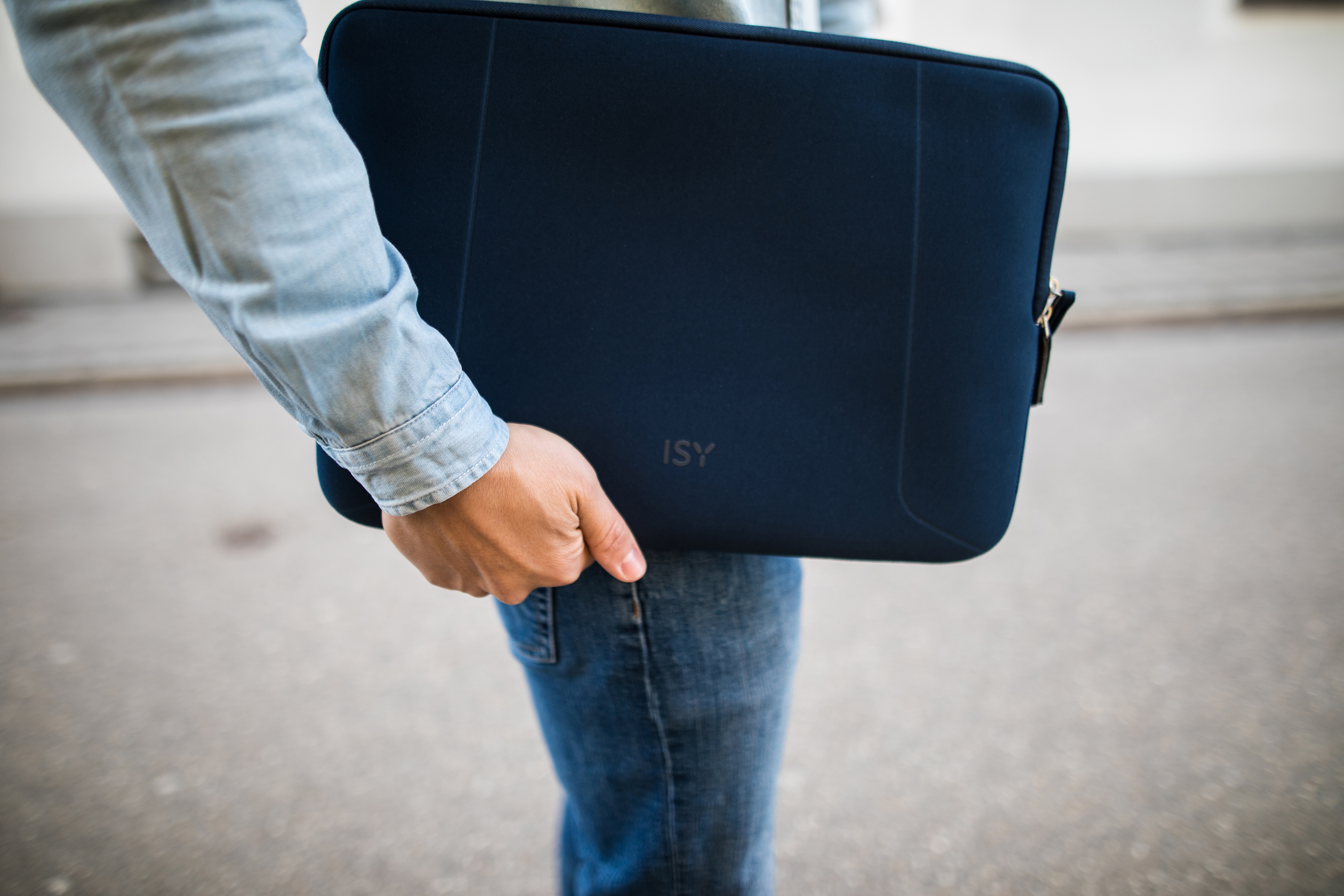 Person hält eine marineblaue ISY-Laptoptasche und geht draußen. Jeans und Jeanshemd.