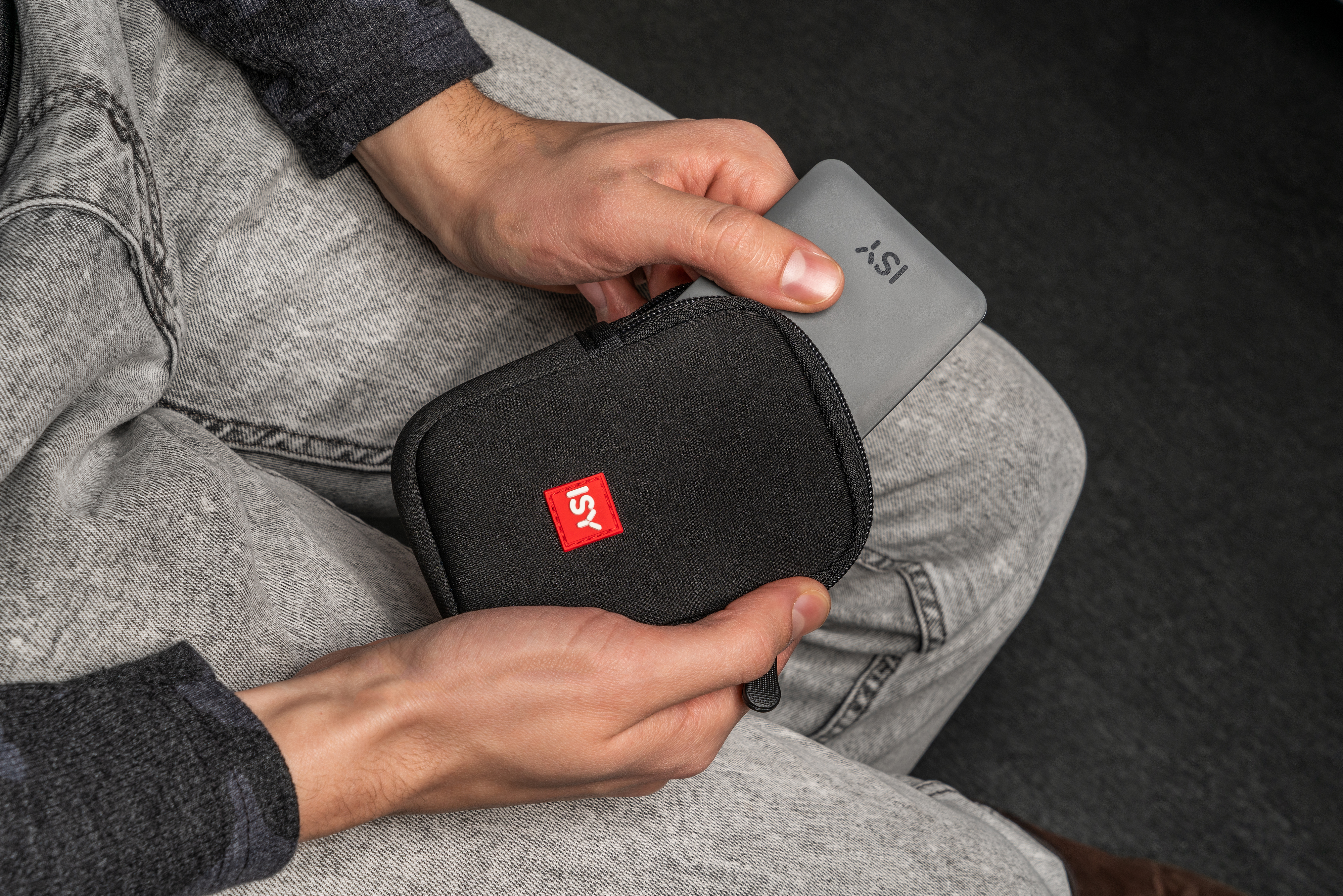Personne tenant un étui noir avec une carte grise étiquetée ISY. La personne est assise sur une surface grise.