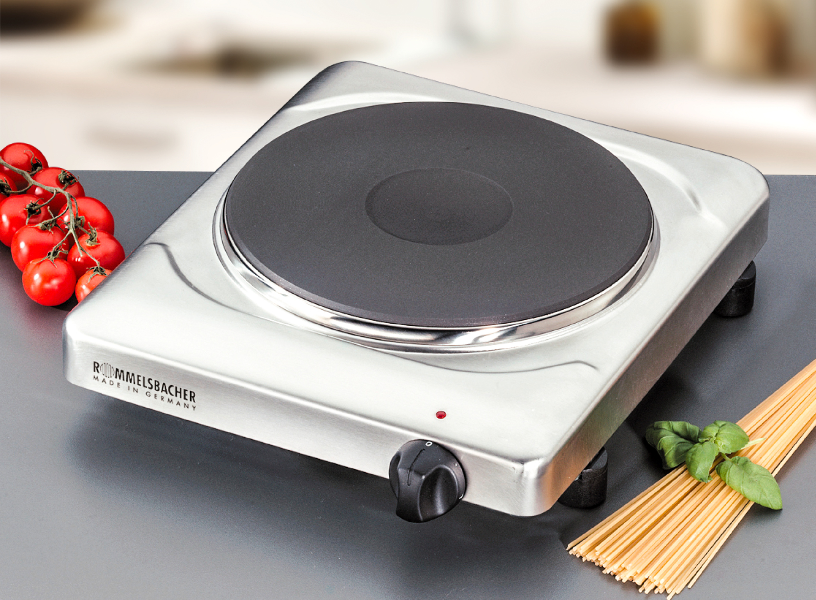 Eine silberne elektrische Kochplatte mit schwarzem Heizelement, Tomaten und Spaghetti.