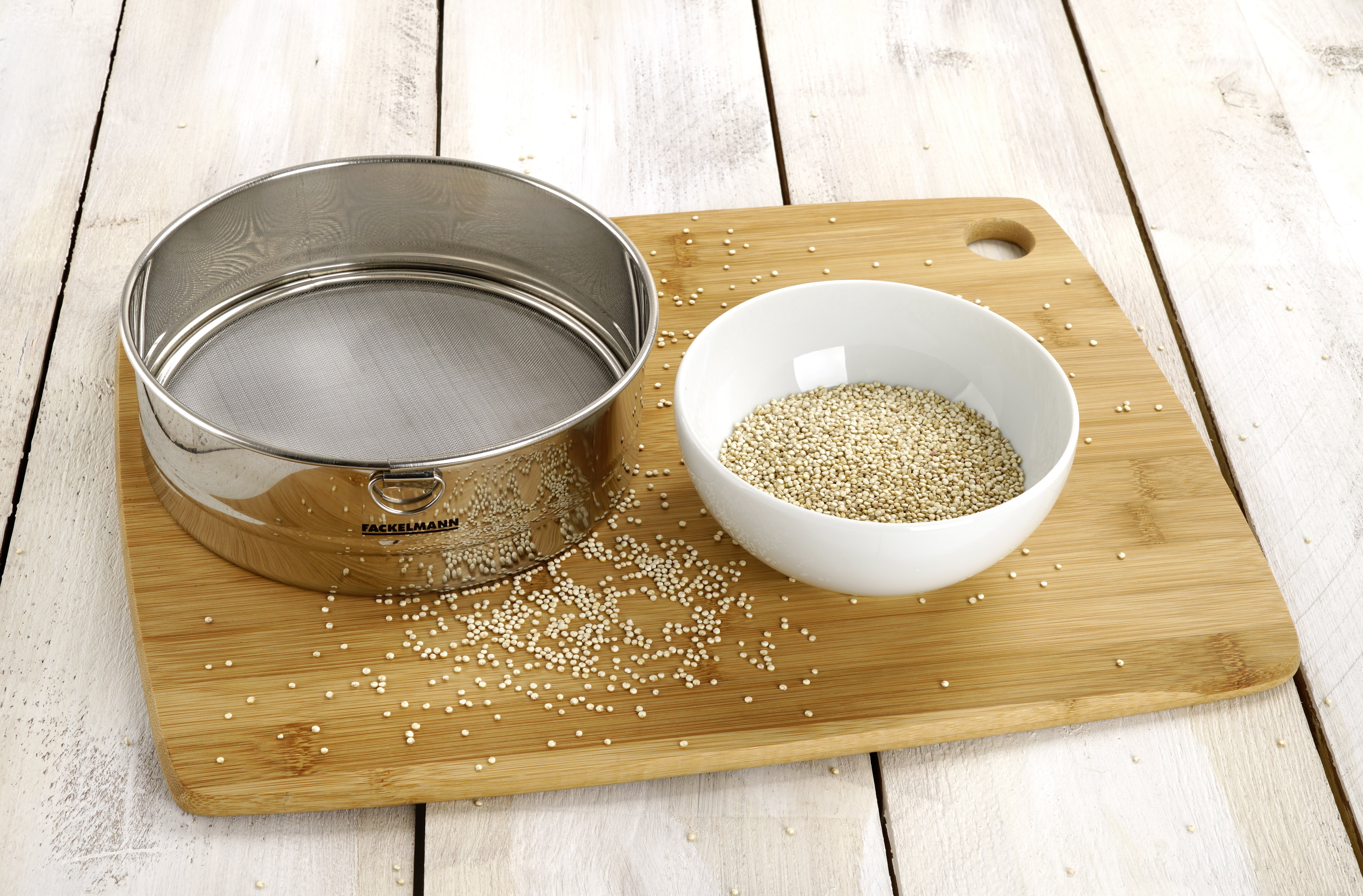 Ein Edelstahlsieb neben einer Schüssel mit Quinoa auf einem Holzschneidebrett.