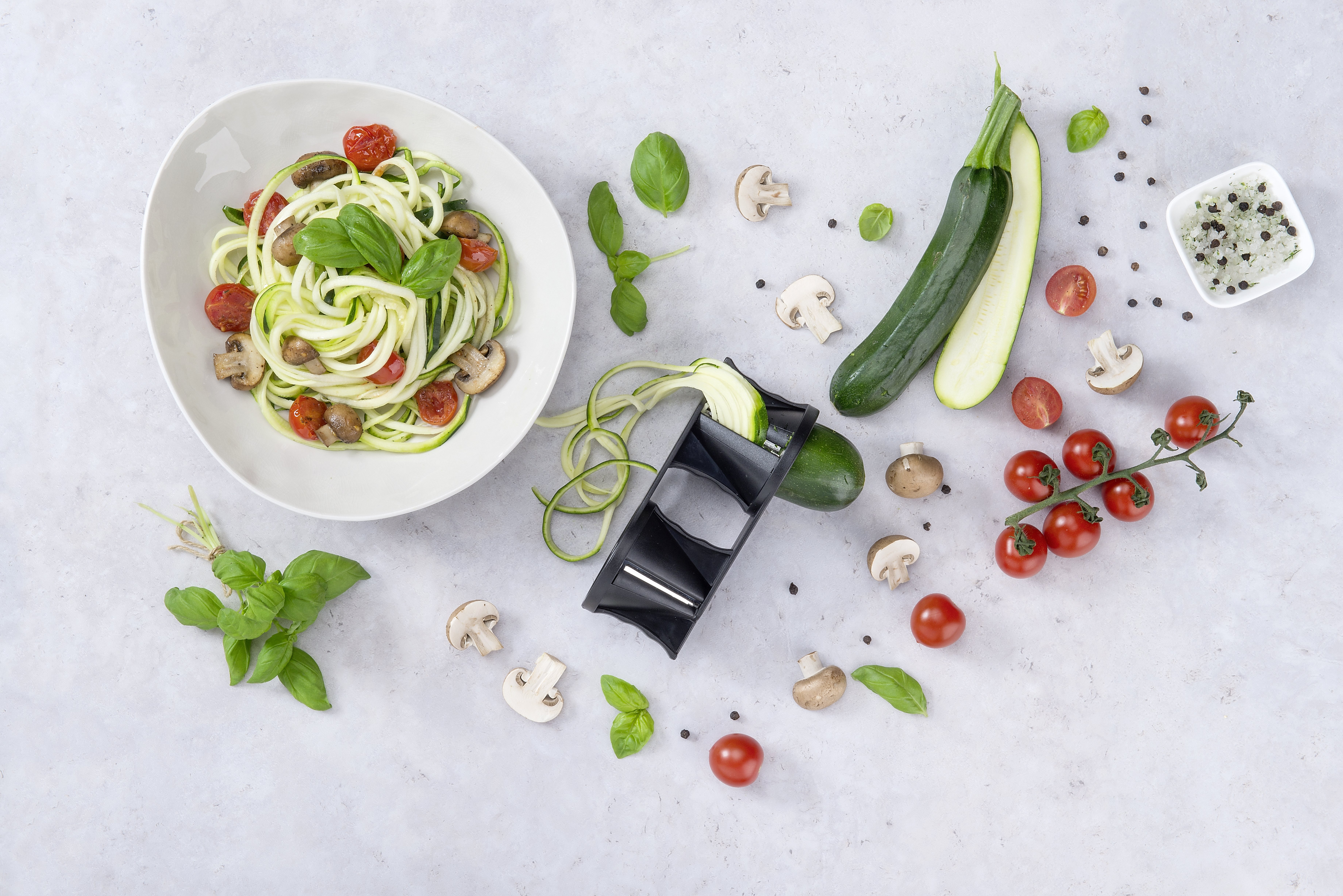 Zucchin-Nudeln mit Tomaten und Pilzen in einer weißen Schüssel, mit Zutaten drum herum. Grauer Hintergrund.