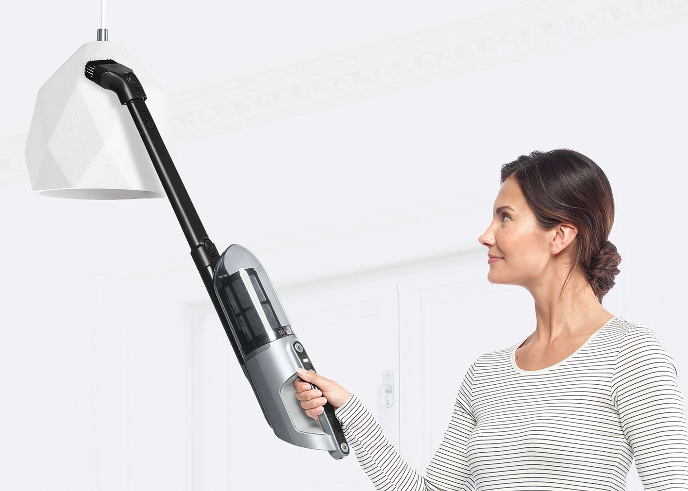 Egy nő egy lámpát tisztít porszívóval. A porszívónak hosszú toldaléka van.