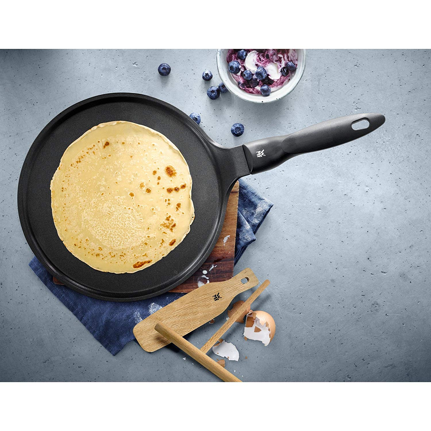 Pfannkuchen in einer schwarzen Pfanne mit Griff auf einem Holzbrett. Blaues Tuch, Werkzeuge.