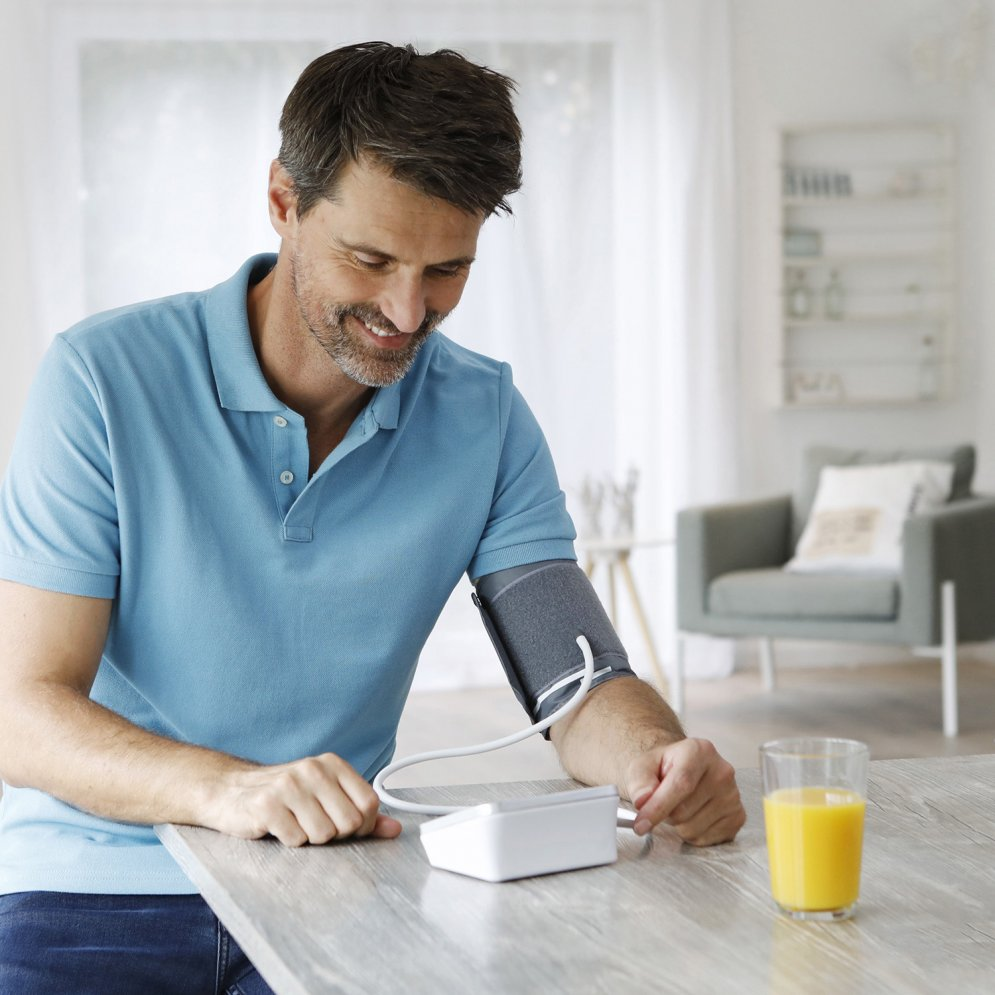 Man checking blood pressure. Sitting at a table with orange juice. White device. Bright background.