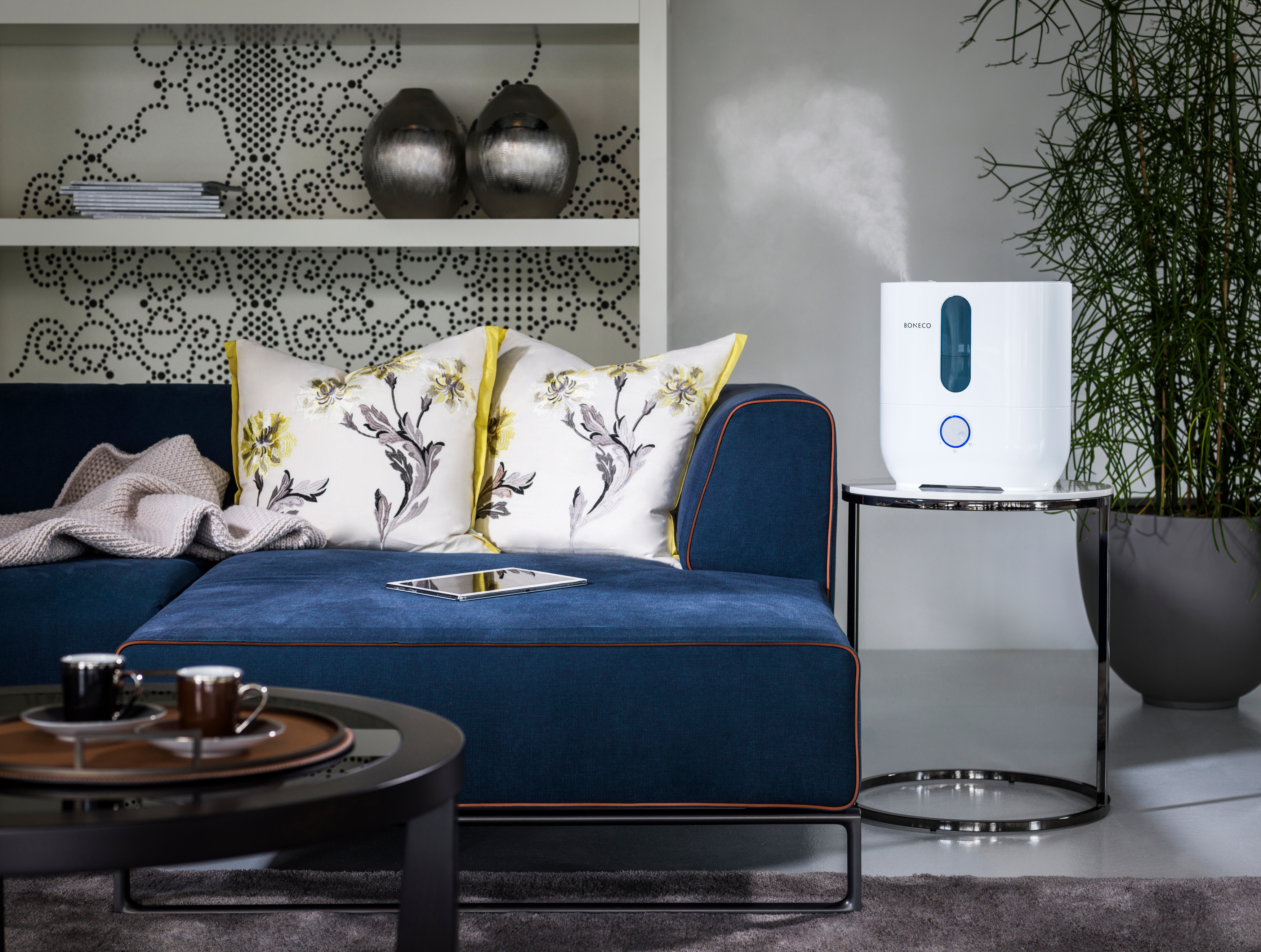 Ein weißer Luftbefeuchter auf einem Tisch neben einem blauen Sofa mit Kissen und einer Pflanze.