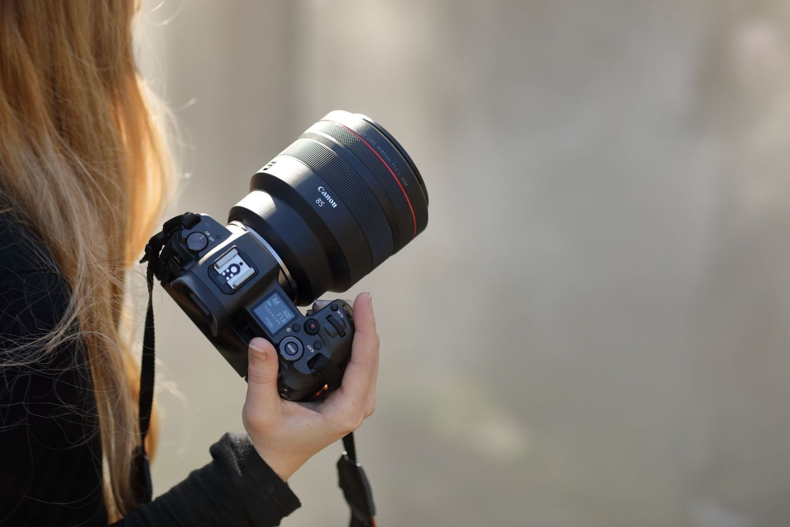Eine Person hält eine Canon-Kamera mit einem 85-mm-Objektiv. Blondes Haar ist sichtbar. Der Hintergrund ist unscharf und neutral.