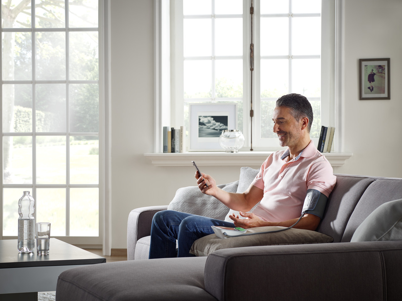 Mann sitzt auf einem Sofa, schaut auf sein Handy, trägt ein Blutdruckmessgerät.