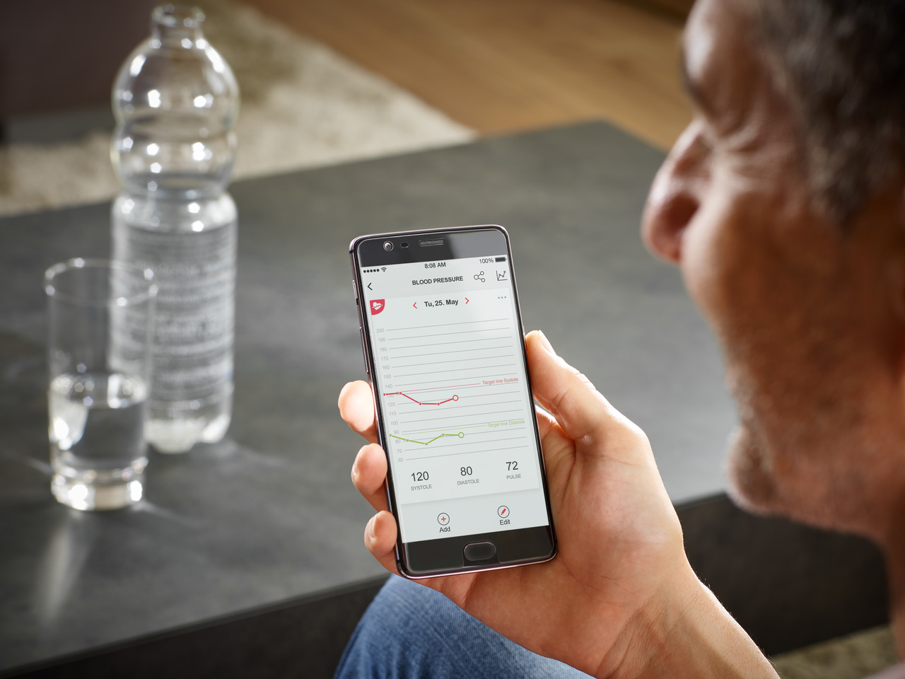 Mann schaut auf ein Smartphone, das eine Blutdrucktabelle anzeigt, eine Wasserflasche und ein Glas sind sichtbar.