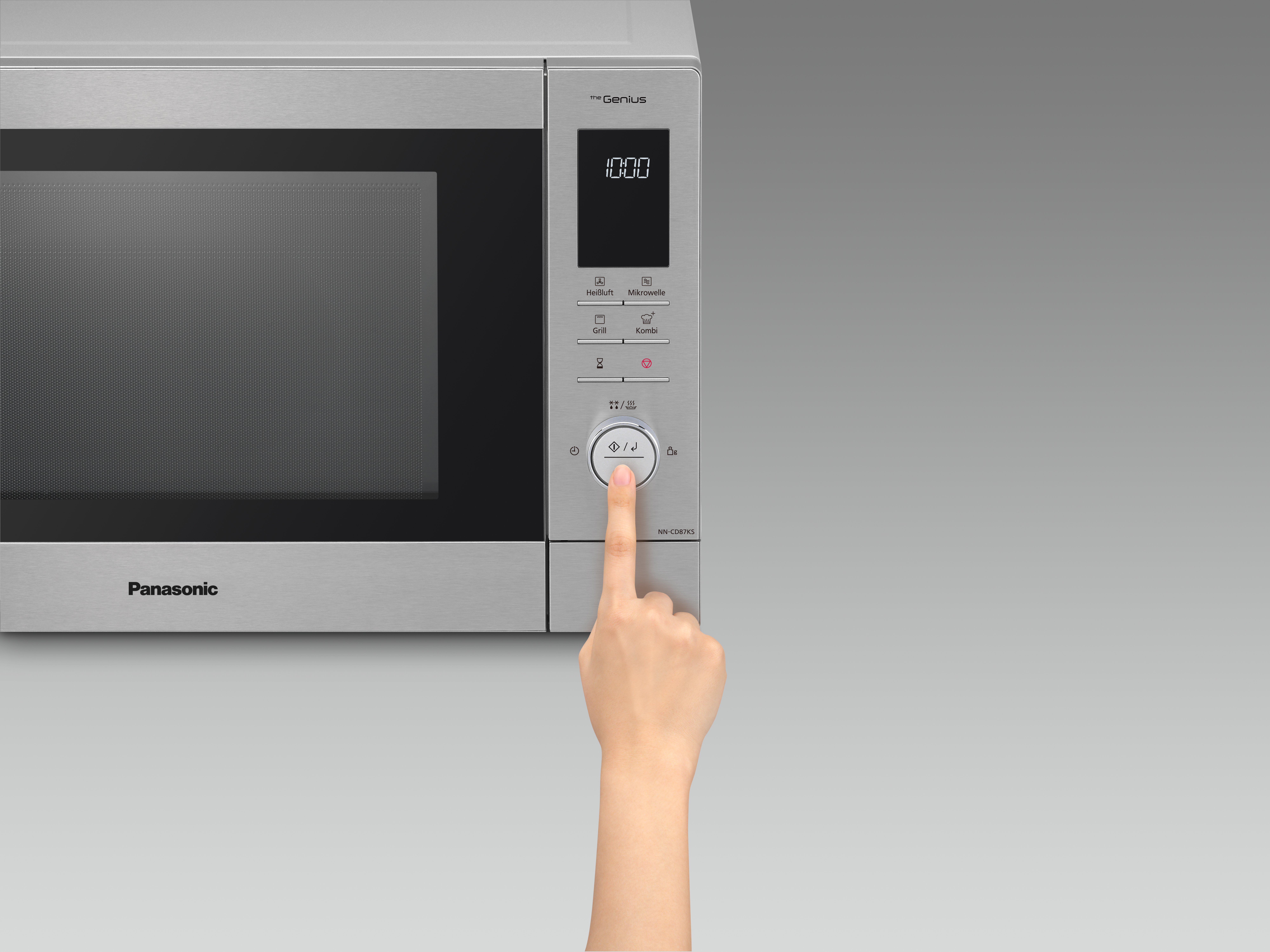 Eine Hand drückt eine silberne Mikrowellentaste. Die Mikrowelle ist silberfarben mit Digitalanzeige.