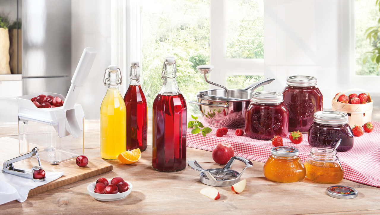 Eine Küchenszene mit Gläsern mit Marmelade, Flaschen mit Saft und Obst auf einer Holztheke. Eine Obstpresse ist sichtbar.