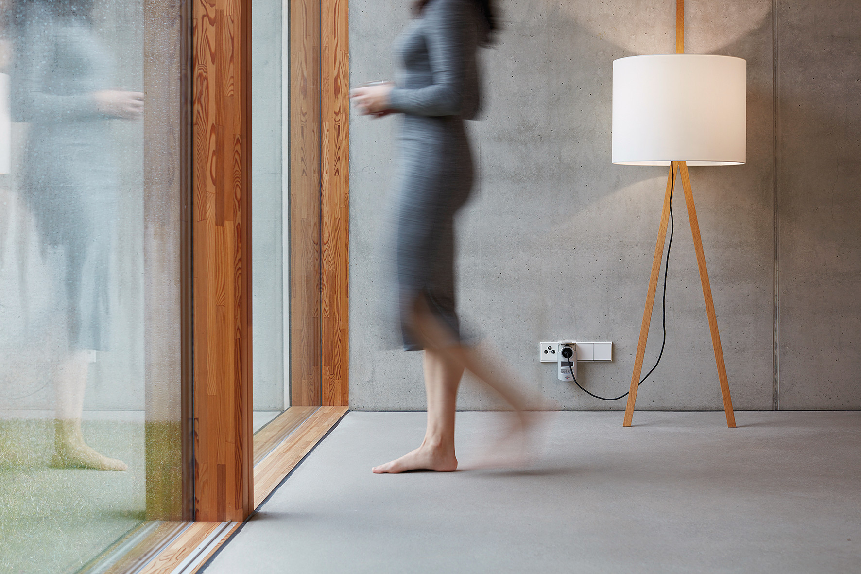 Ein verschwommenes Bild einer Frau, die an einem Fenster vorbeigeht. Eine Lampe ist rechts von ihr. Betonwand.