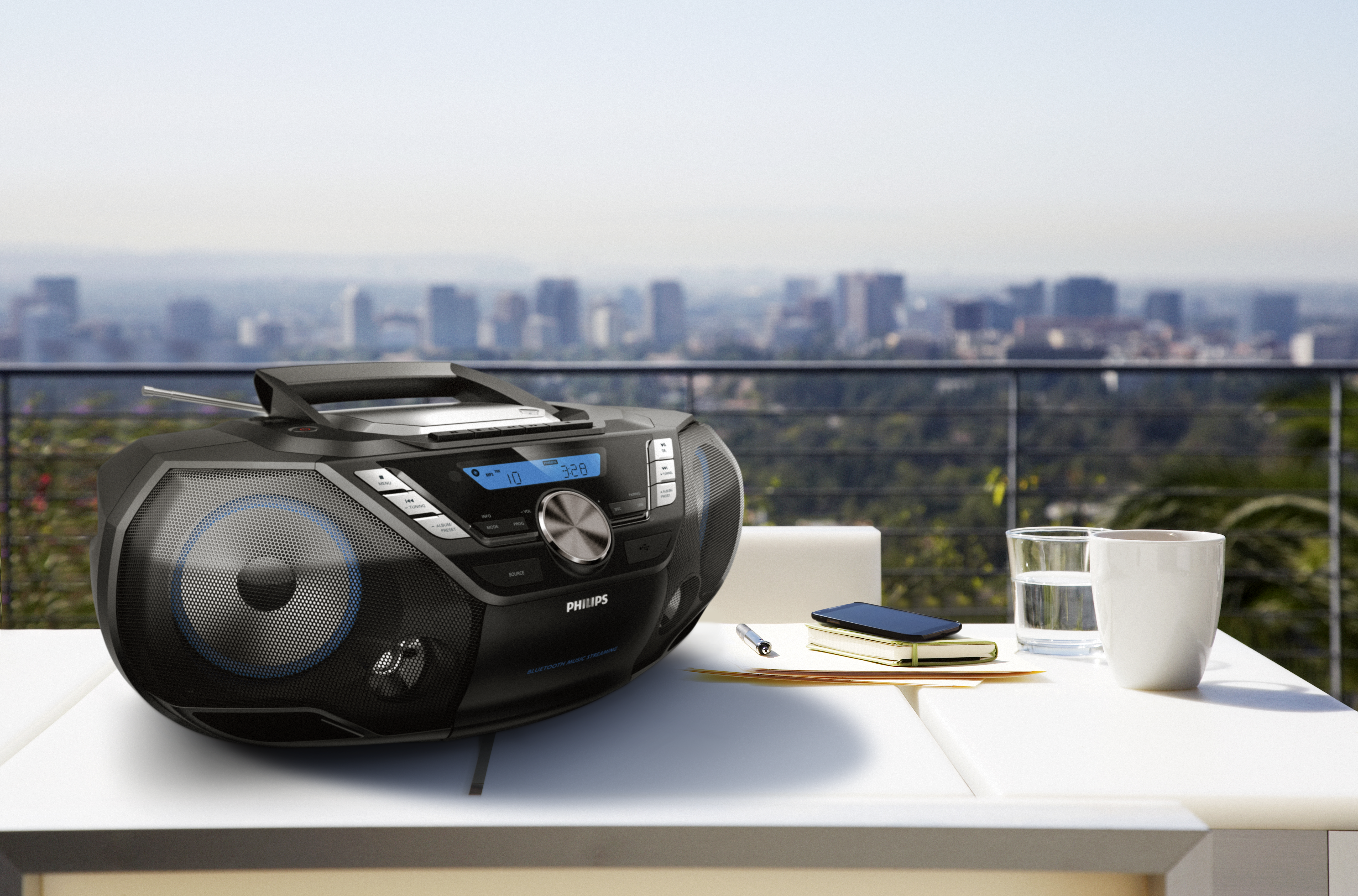 Schwarze Boombox auf weißem Tisch. Stadt im Hintergrund. Glas Wasser und Telefon daneben. Sonniger Tag.