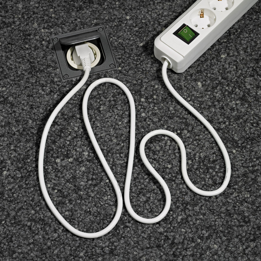 Ein weißes Elektrokabel und ein Stecker, die an einer Steckdosenleiste auf einem Teppich angeschlossen sind.