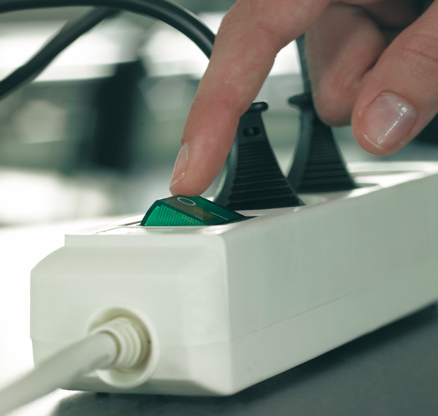 Eine Hand drückt den grünen Netzschalter an einer weißen Steckdosenleiste. Schwarze Kabel und Stecker sind sichtbar.