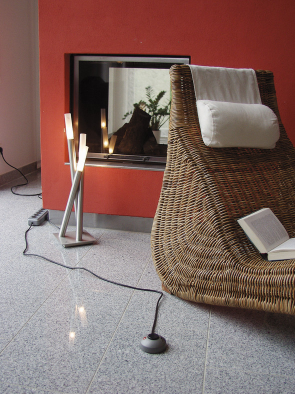 Wohnzimmer mit Korbstuhl, Lampe und Kamin. Ein Buch ist auf dem Stuhl geöffnet.