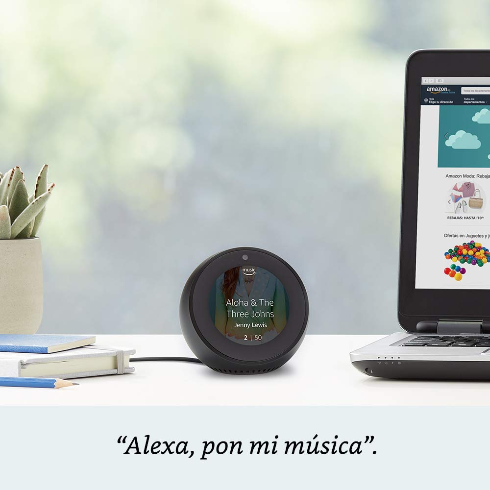Pantalla de altavoz inteligente negro muestra info de música, portátil a la derecha con sitio web Amazon abierto.