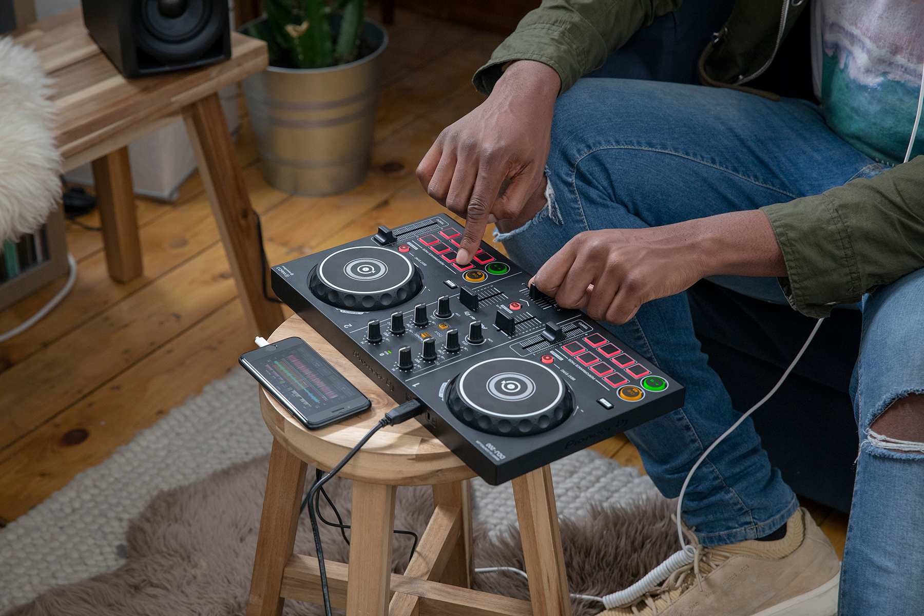 A person is DJing with a Pioneer DJ controller on a wooden stool. A phone sits beside it.