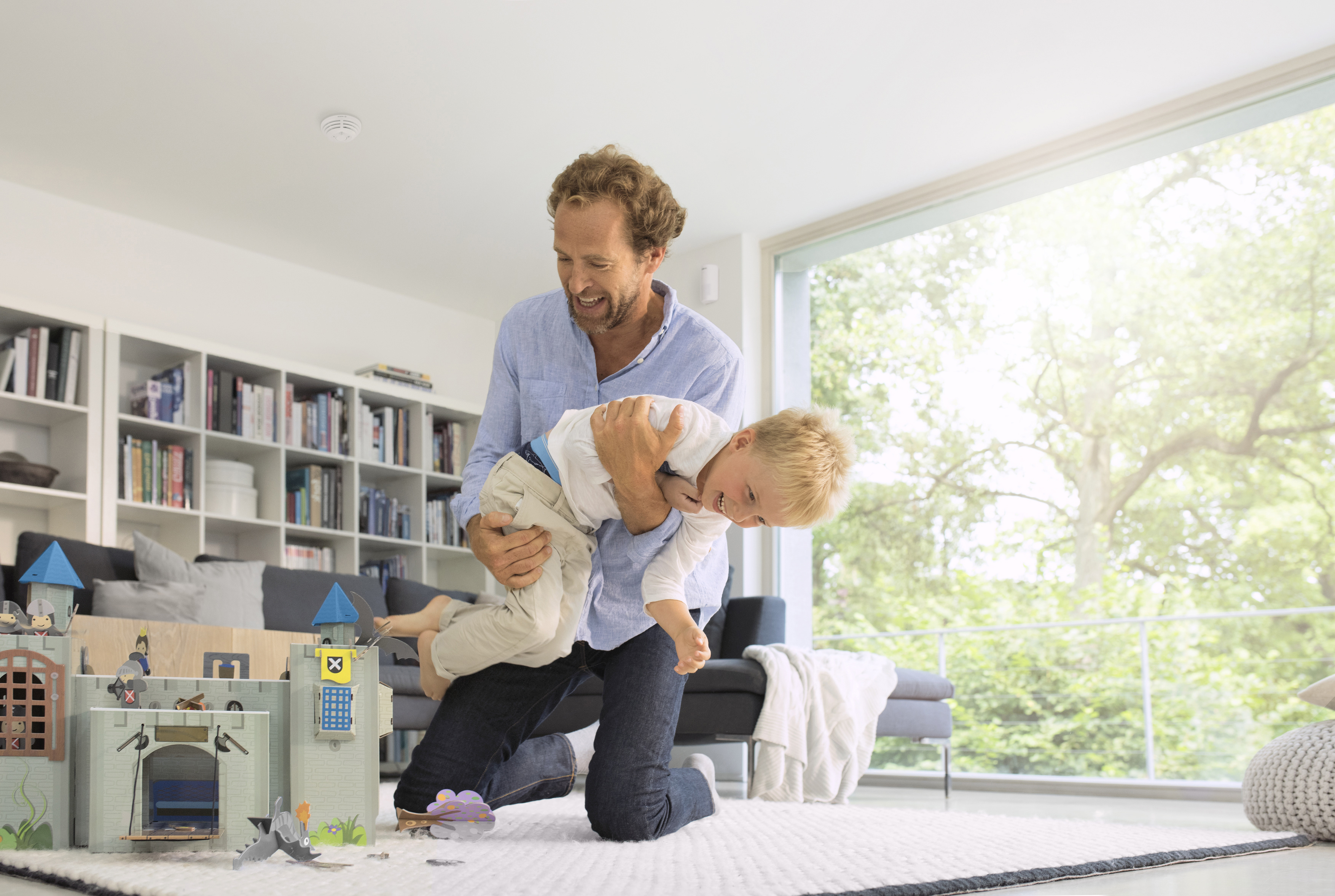 Ein Vater hält seinen Sohn spielerisch im Wohnzimmer, in der Nähe ein Spielschloss.