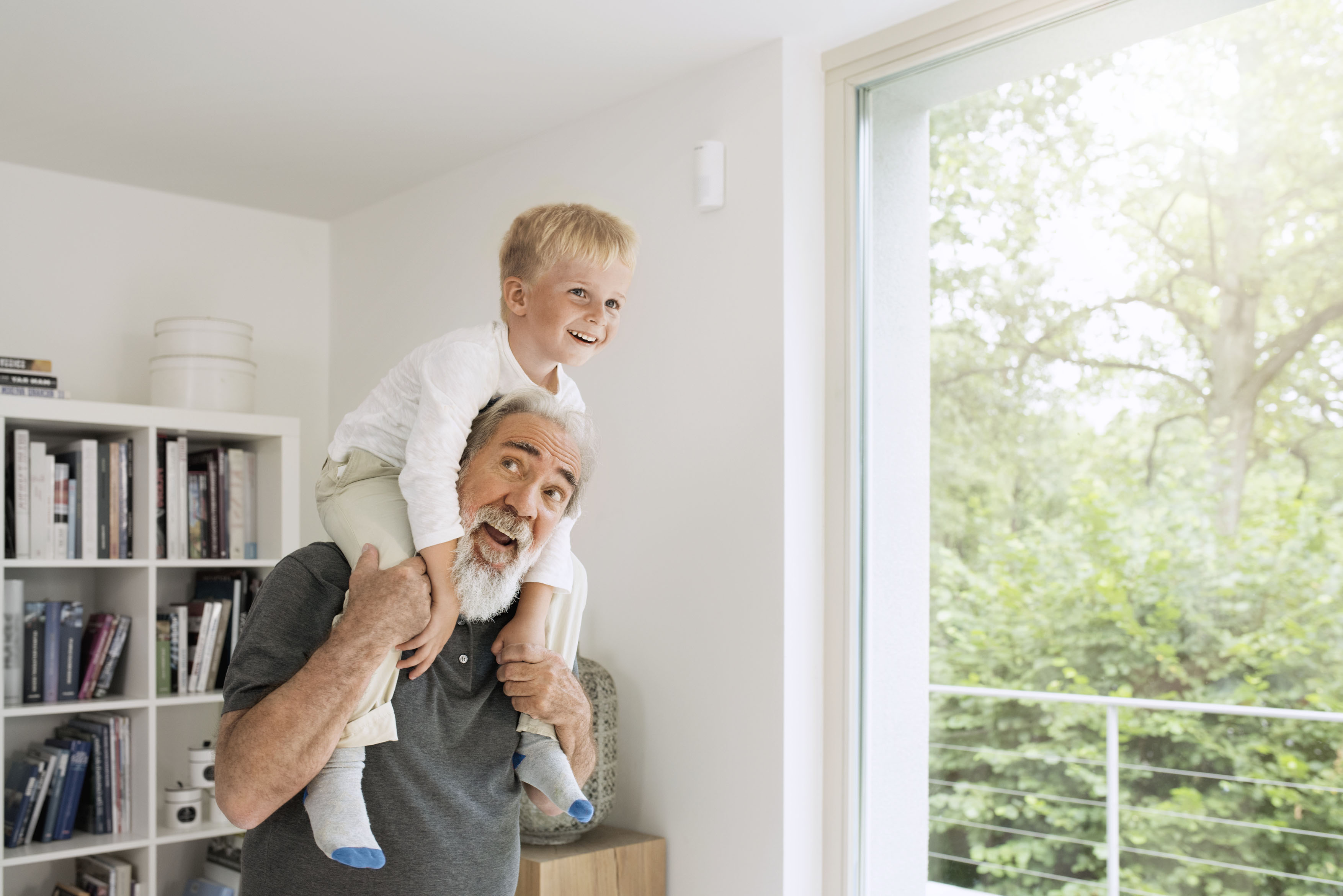 Großvater mit Kind auf den Schultern, lächelnd in einem Raum mit Fenster.