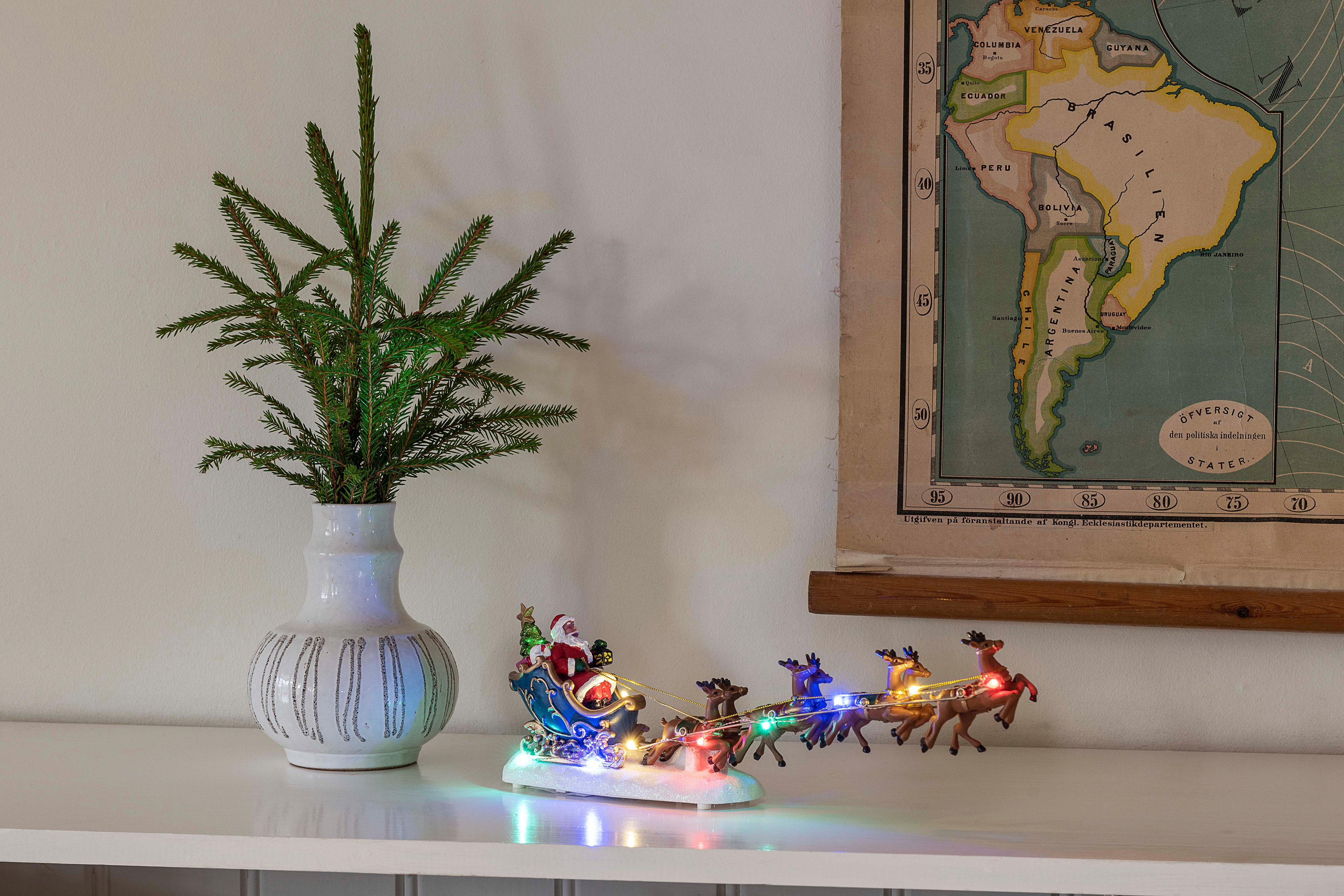 Weihnachtsmann im Schlitten mit Rentieren, Baum in Vase und Vintage-Karte auf weißem Regal.