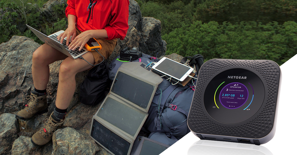 Person mit Laptop, Solarpanel, Tablet und tragbarem WLAN-Gerät auf einem Berg. Hintergrund ist ein grüner Wald.