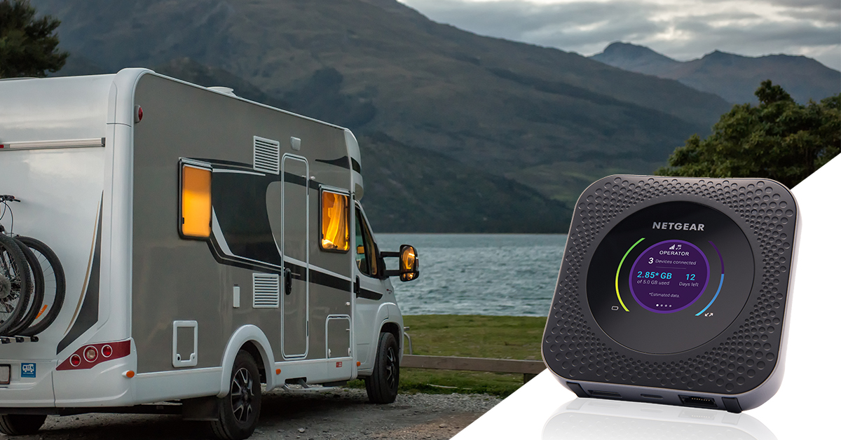Wohnmobil am See, Netgear WLAN-Gerät im Vordergrund. Abendlicht in den Fenstern des Wohnmobils. Berg im Hintergrund.