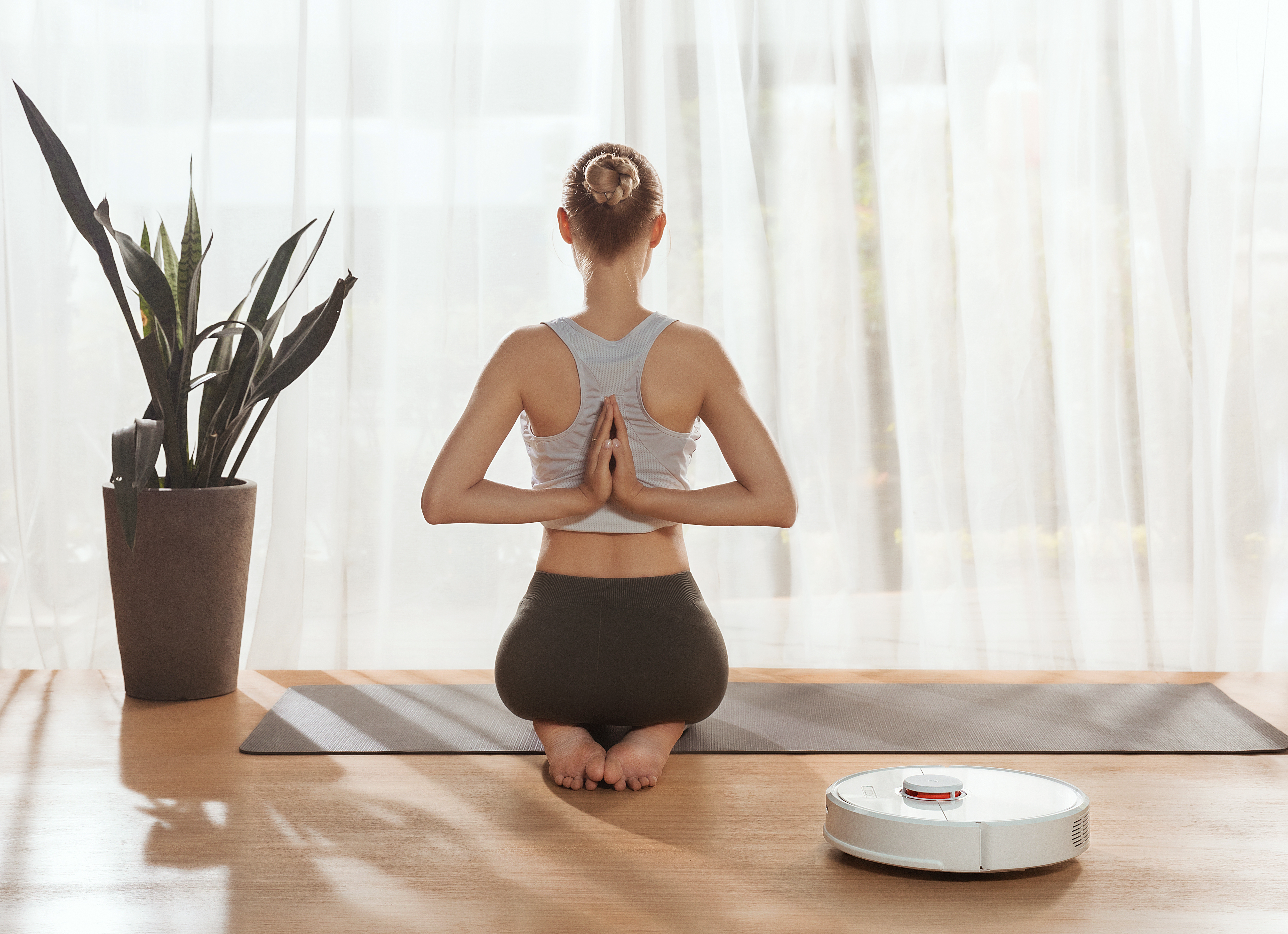Frau in Yogahaltung mit Händen hinter dem Rücken auf Matte. Pflanze und Roboter-Staubsauger in hellem Raum.