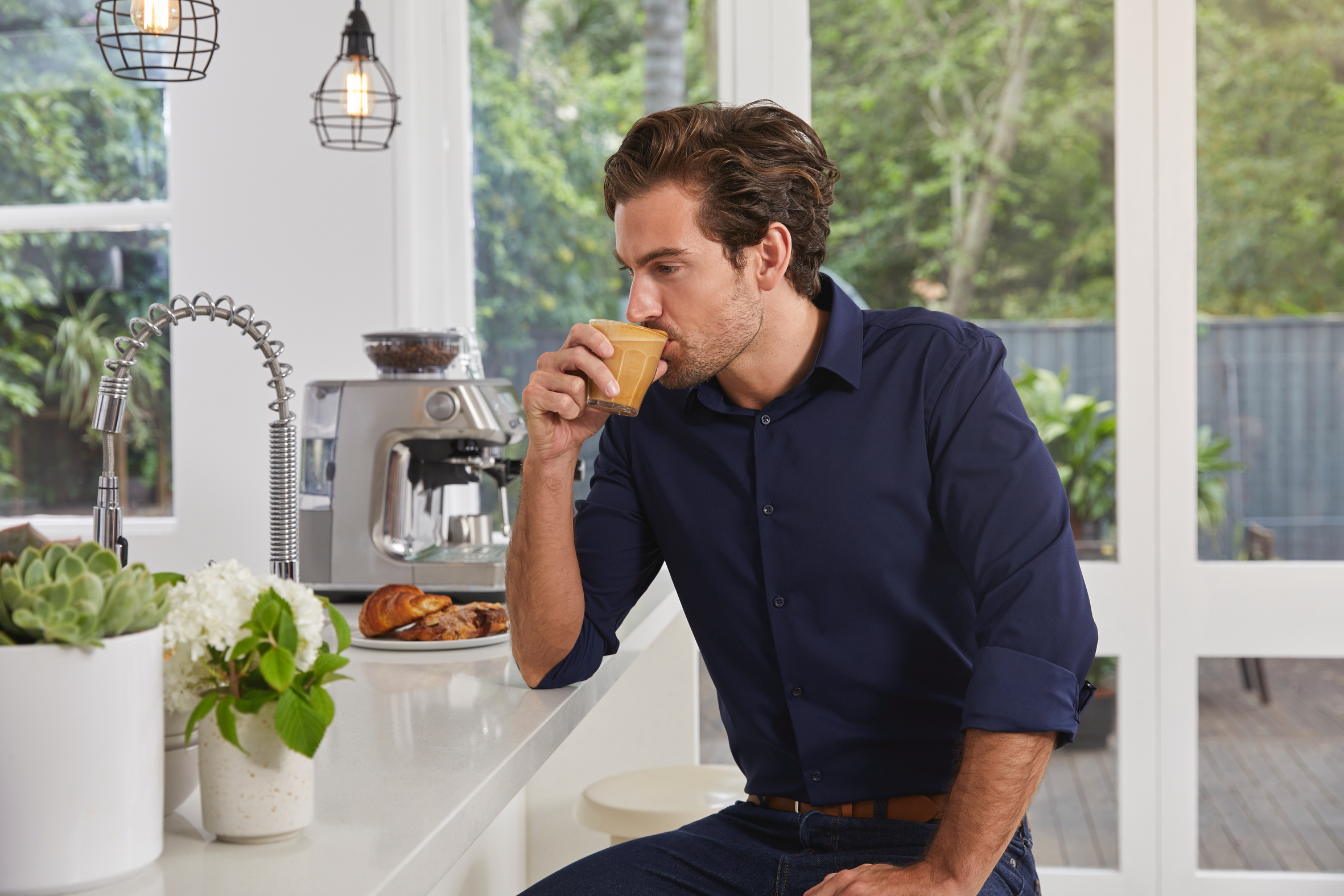 Ein Mann trinkt Kaffee in einer Küche. In der Nähe befinden sich eine Kaffeemaschine und Gebäck.