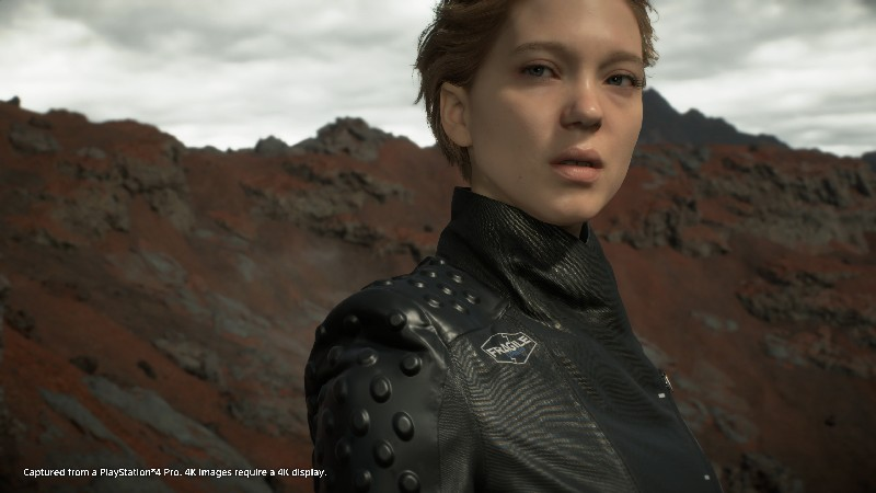 Una mujer con una chaqueta negra mira al espectador. El fondo es un paisaje rocoso y rojo. El juego es Death Stranding.