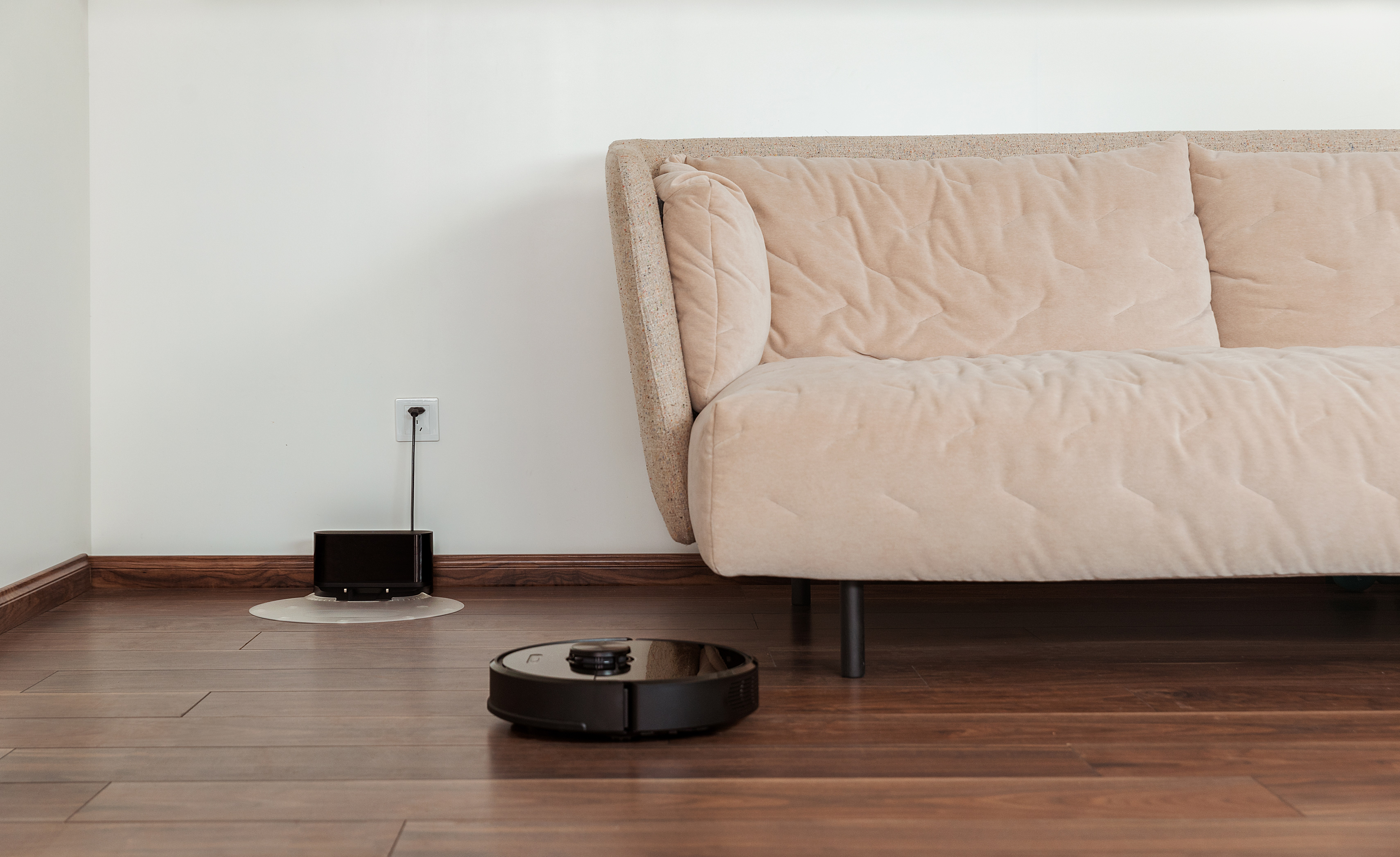 Ein schwarzer Roboter-Staubsauger auf einem Holzboden. Rechts ein beiges Sofa. Eine Ladestation links. Weiße Wand.