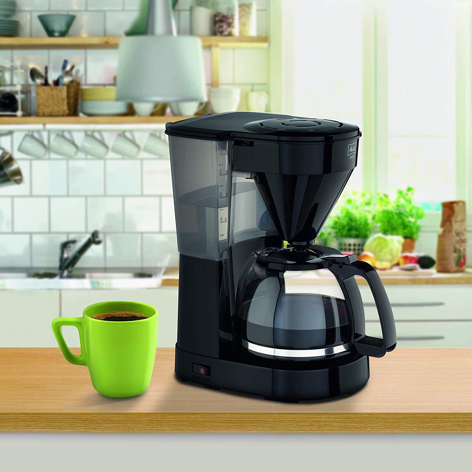Macchina da caffè nera con una tazza verde. Sfondo cucina con mobili bianchi e piante verdi.