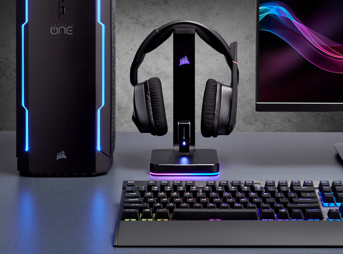 Gaming setup: PC, headphones on a stand, keyboard with lights. Grey desk and dark background.