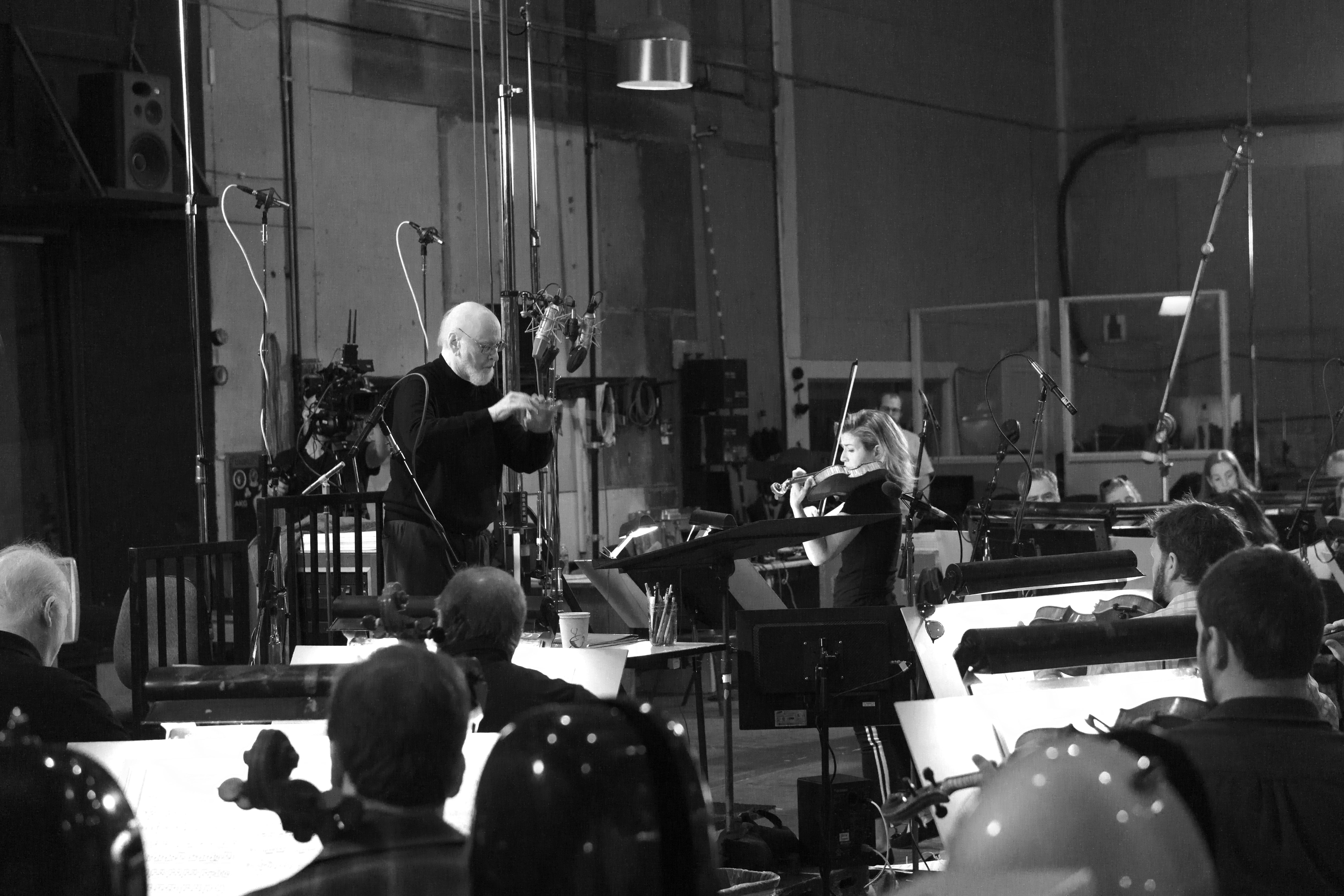 Ein Dirigent leitet ein Orchester, eine Geigerin spielt, und Musiker lesen Noten in einem Studio.