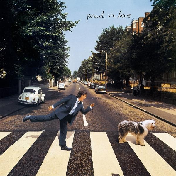 Paul McCartney auf einem Zebrastreifen, mit einem Hund. Autos und Bäume im Hintergrund, eine Straßenszene.