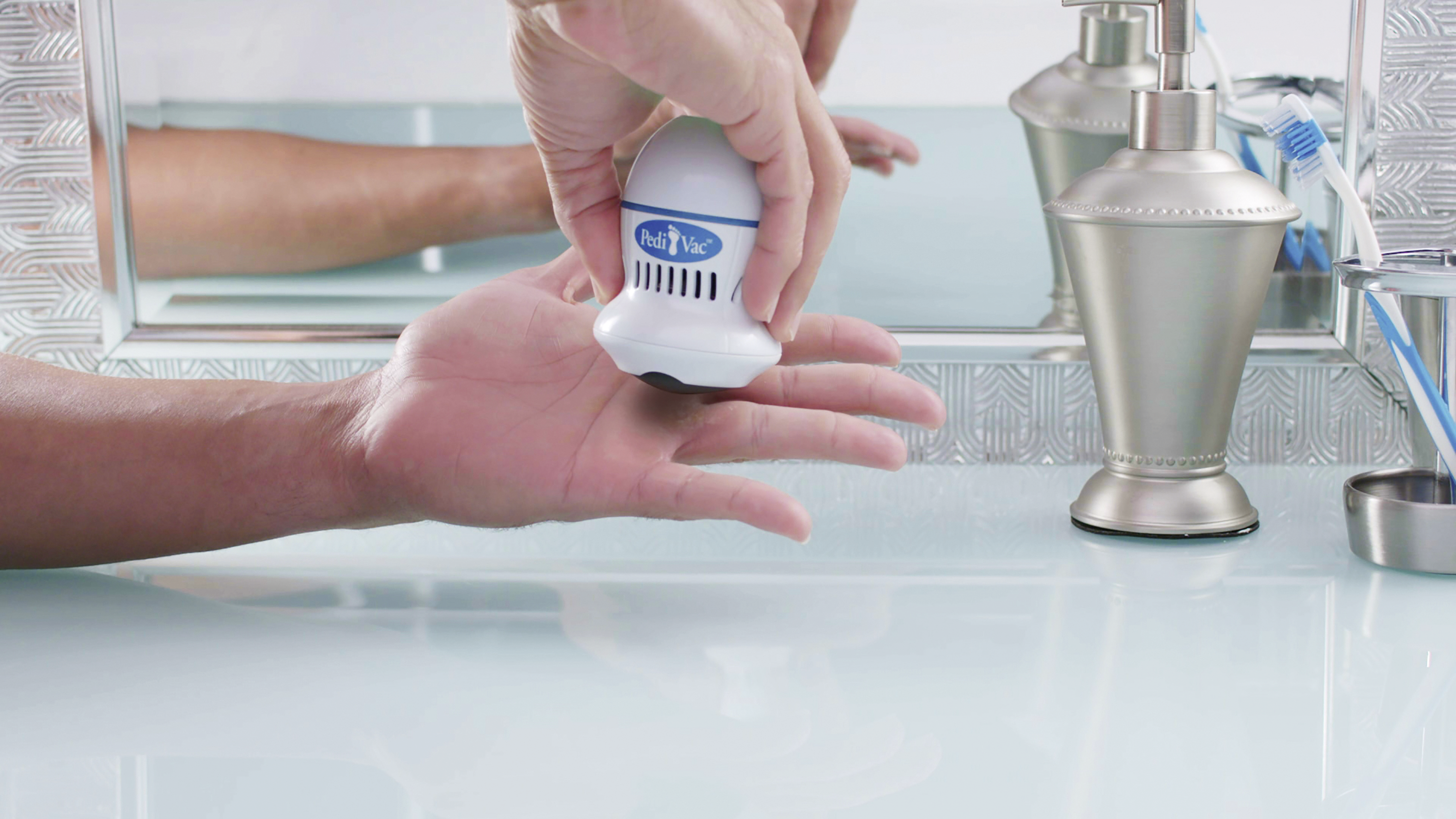 Person benutzt einen weißen Pedi Vac auf der Hand über einem Waschbecken, mit Spiegel dahinter.