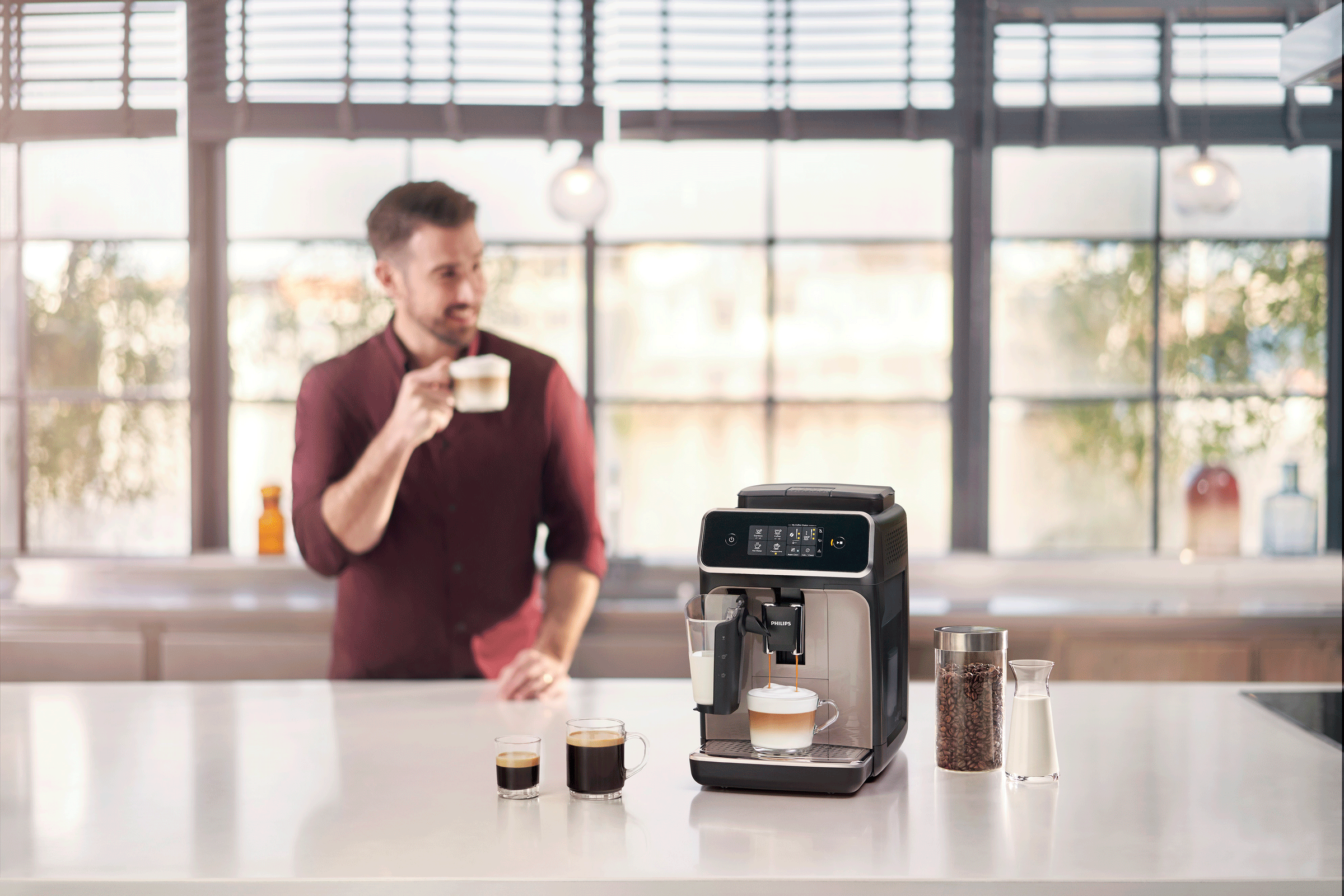 Mann macht Kaffee mit einer Maschine. Tassen und eine Kaffeemühle stehen auf der Theke. Küchenfenster ist sichtbar.