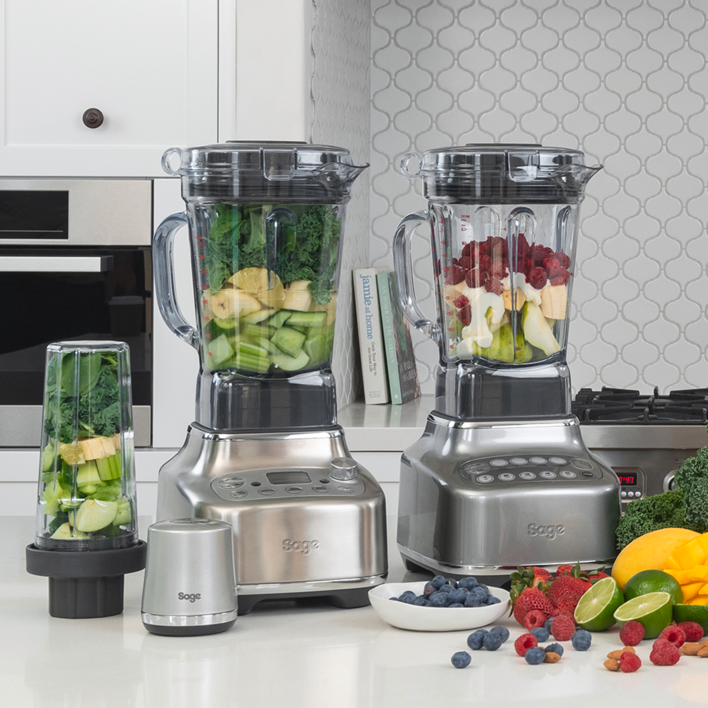 Drei silberne Mixer mit Zutaten auf einer weißen Theke, frisches Obst und Gemüse.