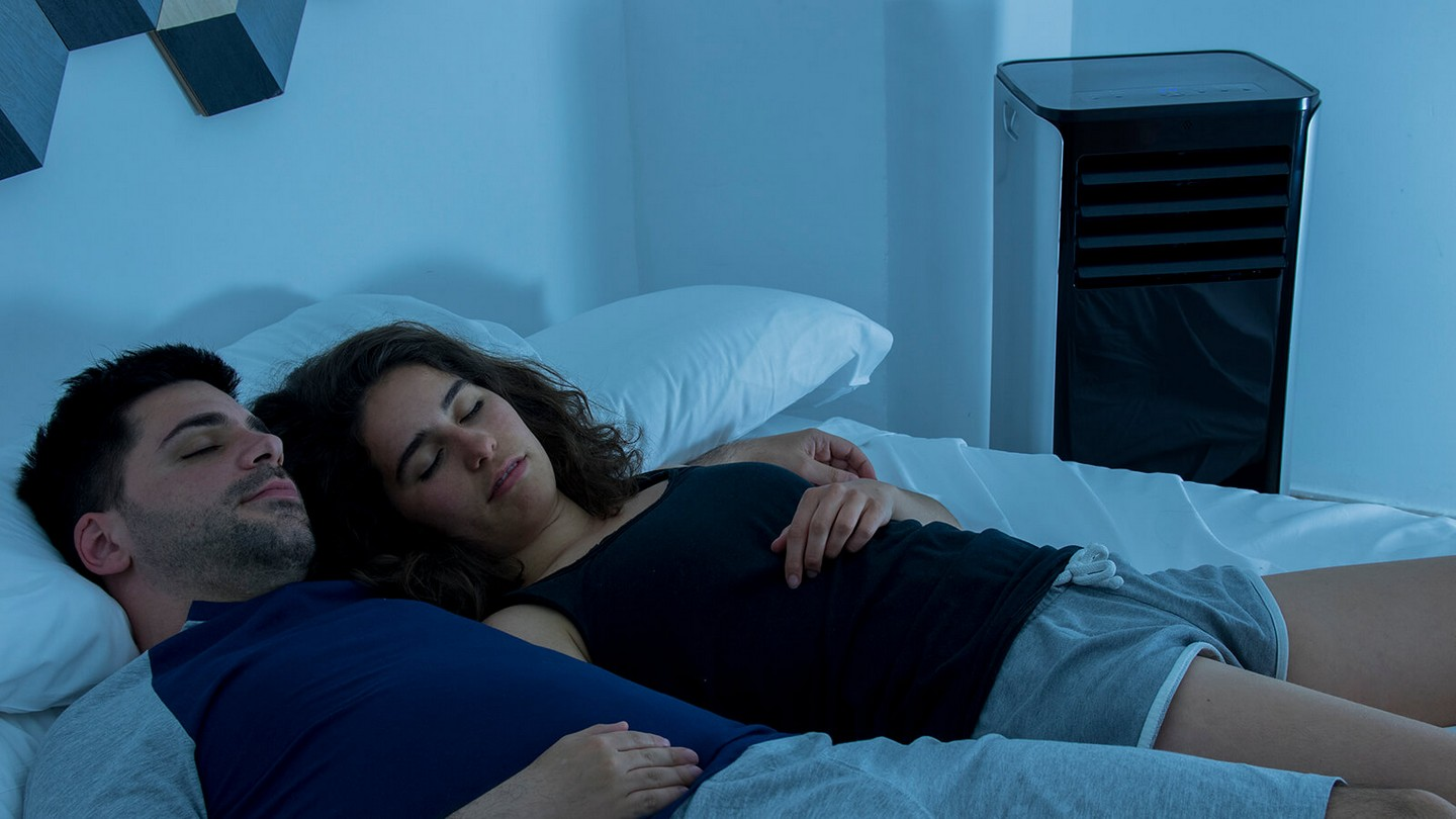 Pareja durmiendo en la cama, abrazados. Habitación oscura, luz azul de un enfriador de aire negro en el fondo.
