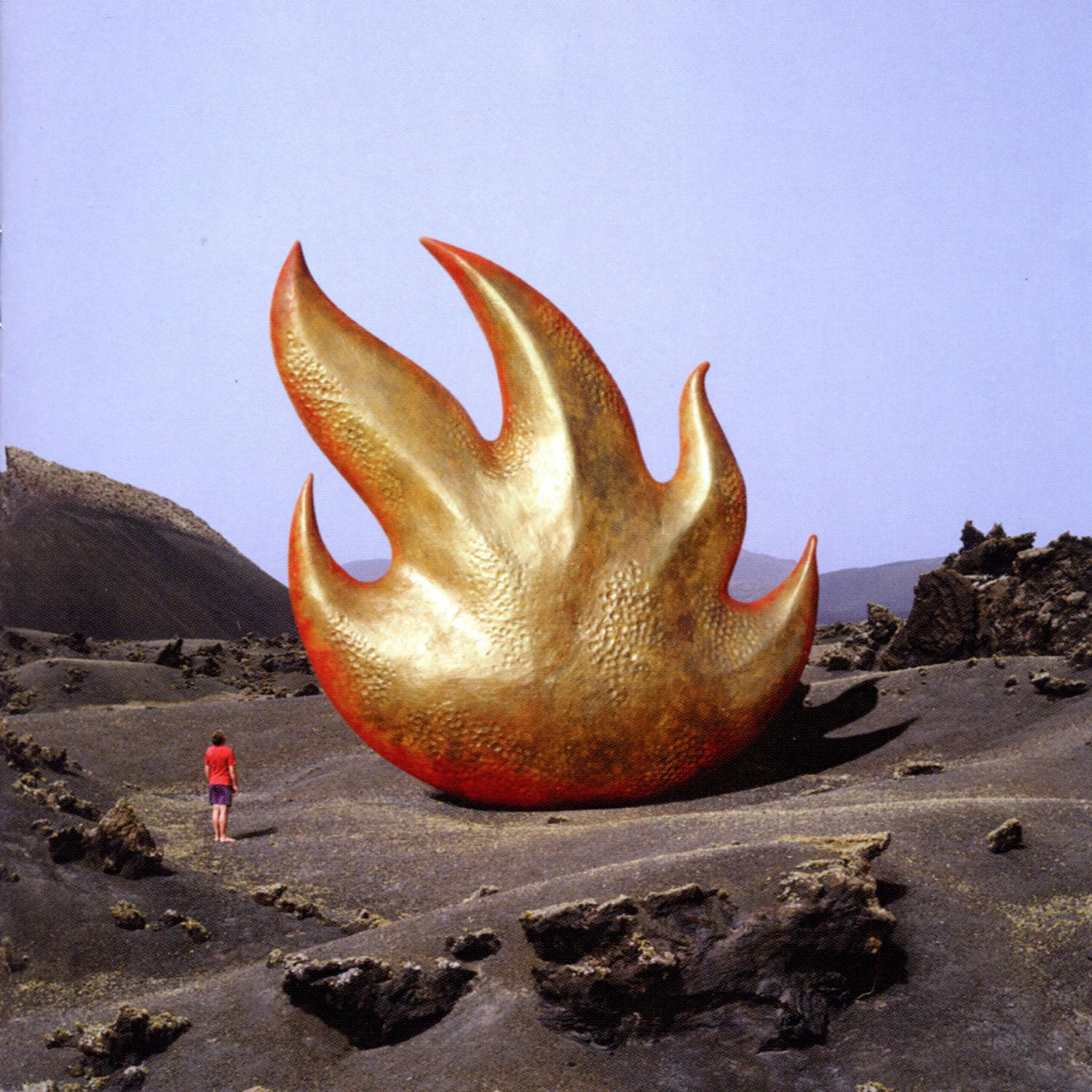 Eine goldene Flammenskulptur in einer vulkanischen Landschaft. Eine Person geht weg. Blauer Himmel.